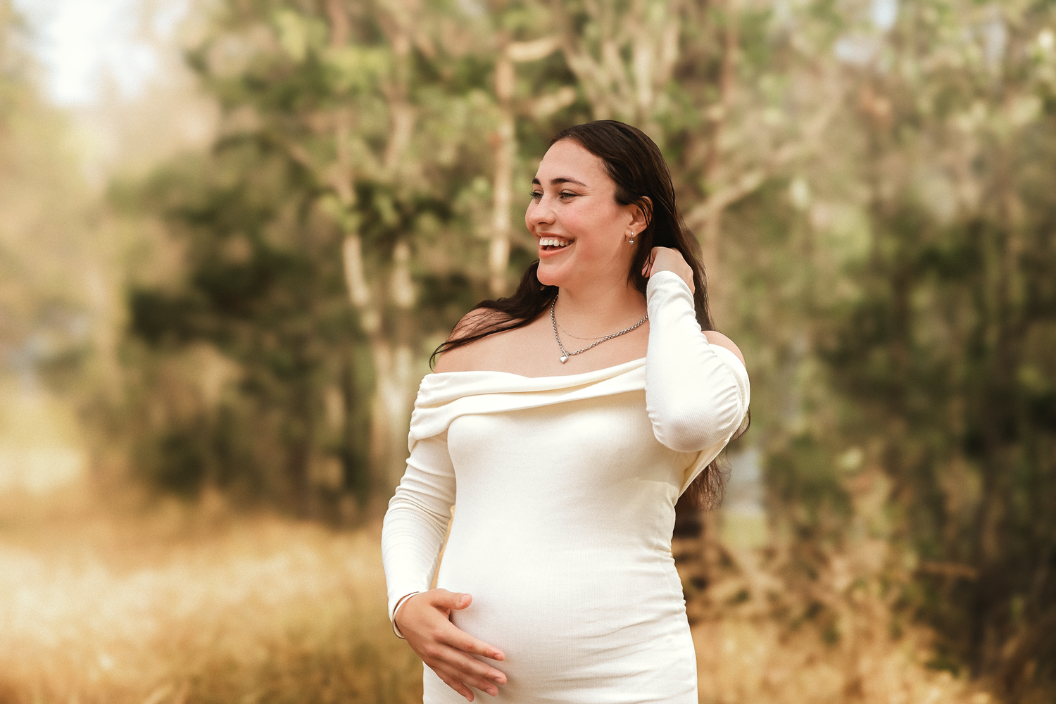 Smiling pregnant woman in a white off-the-shoulder dress cradling her baby bump during an outdoor maternity photoshoot among autumn trees.