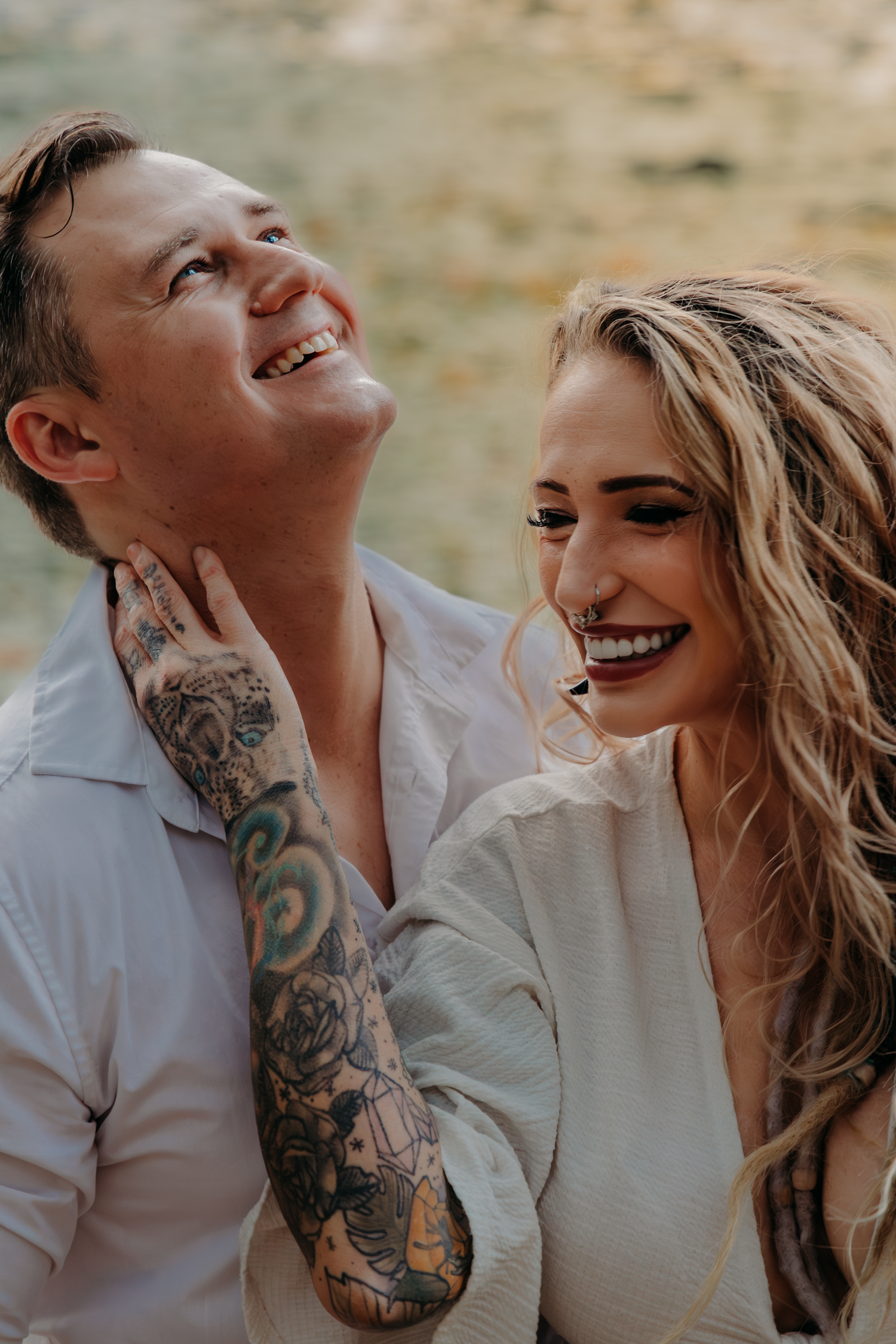 A laughing couple in white shirts embrace outdoors, the woman showing tattooed arms.