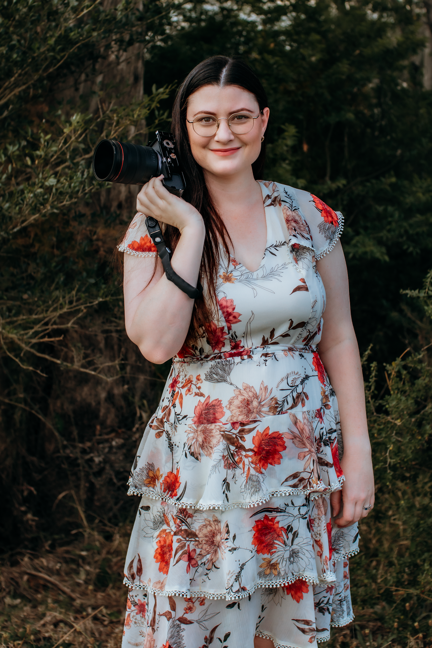 Young woman in floral dress holding a professional camera outdoors, smiling, wearing glasses, surrounded by lush greenery.