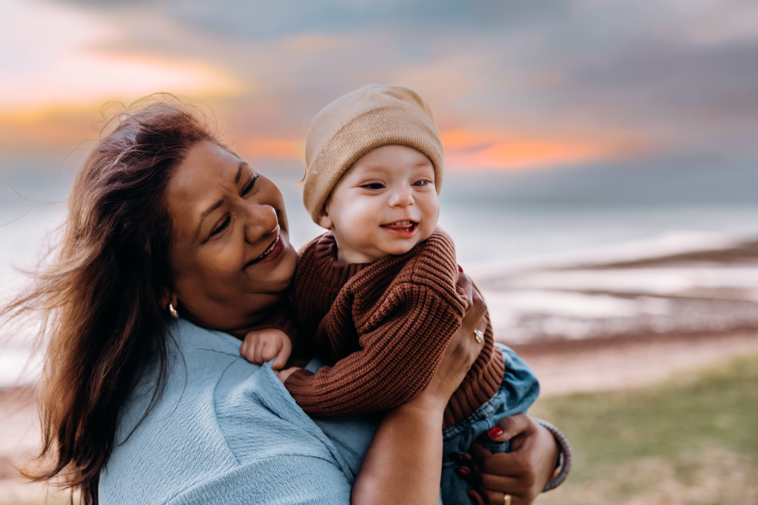 A mother smiles warmly while holding her happy baby outdoors near the ocean at sunset.