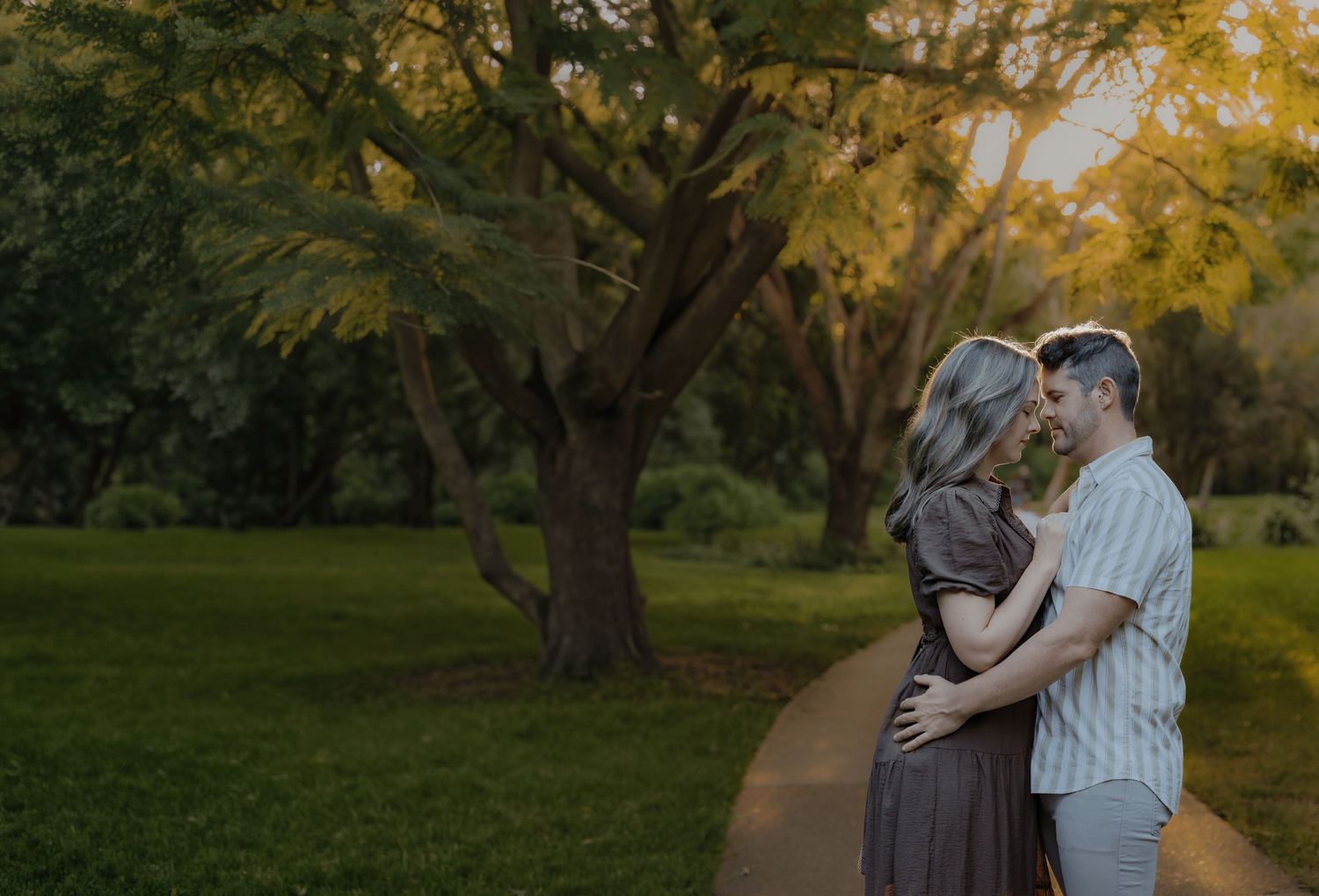 Couple embracing and nearly kissing on a garden path surrounded by lush green trees in warm golden light.