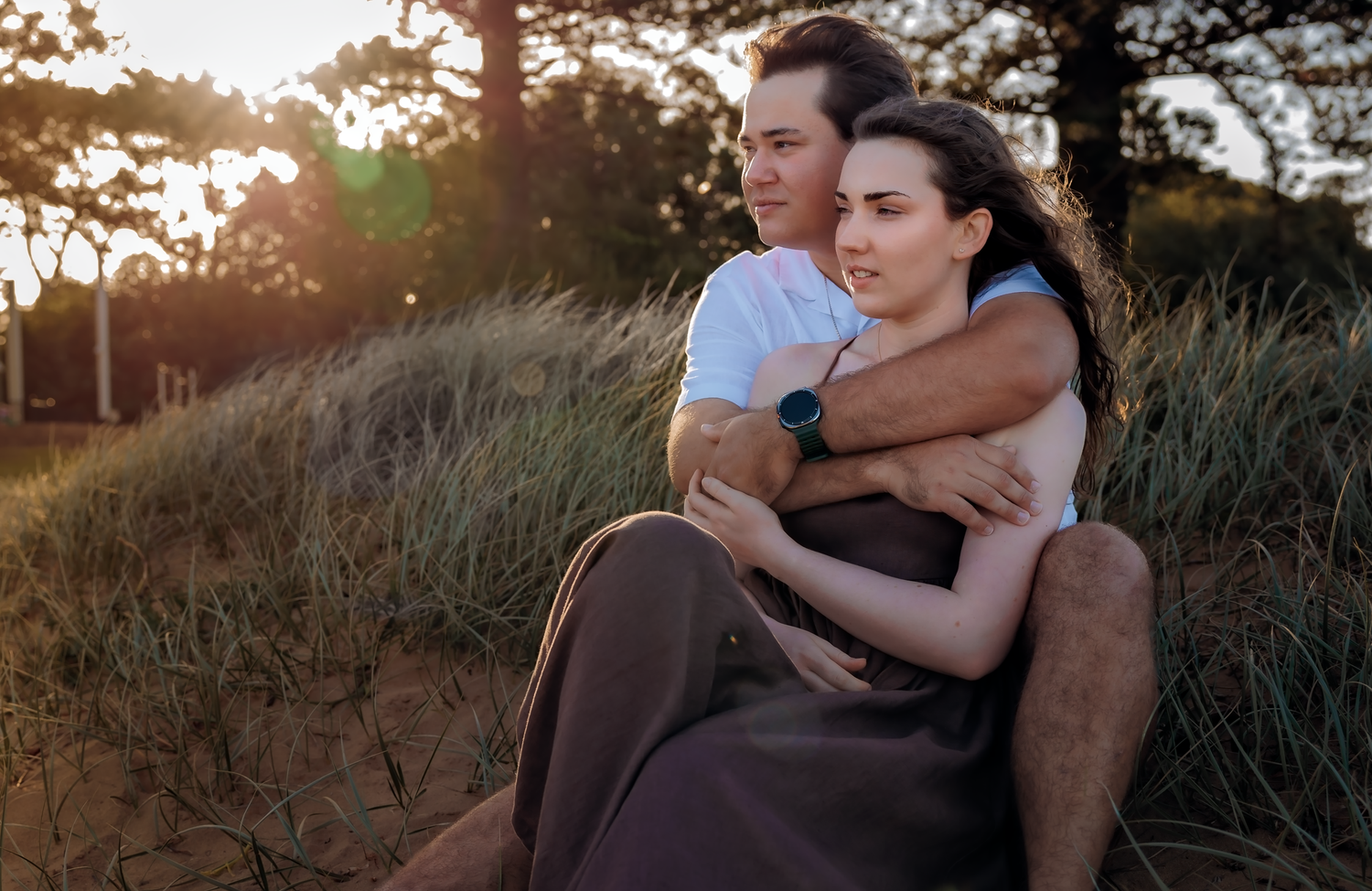Couple embracing on sandy dunes at golden hour, woman in brown skirt leaning back against man in white shirt.