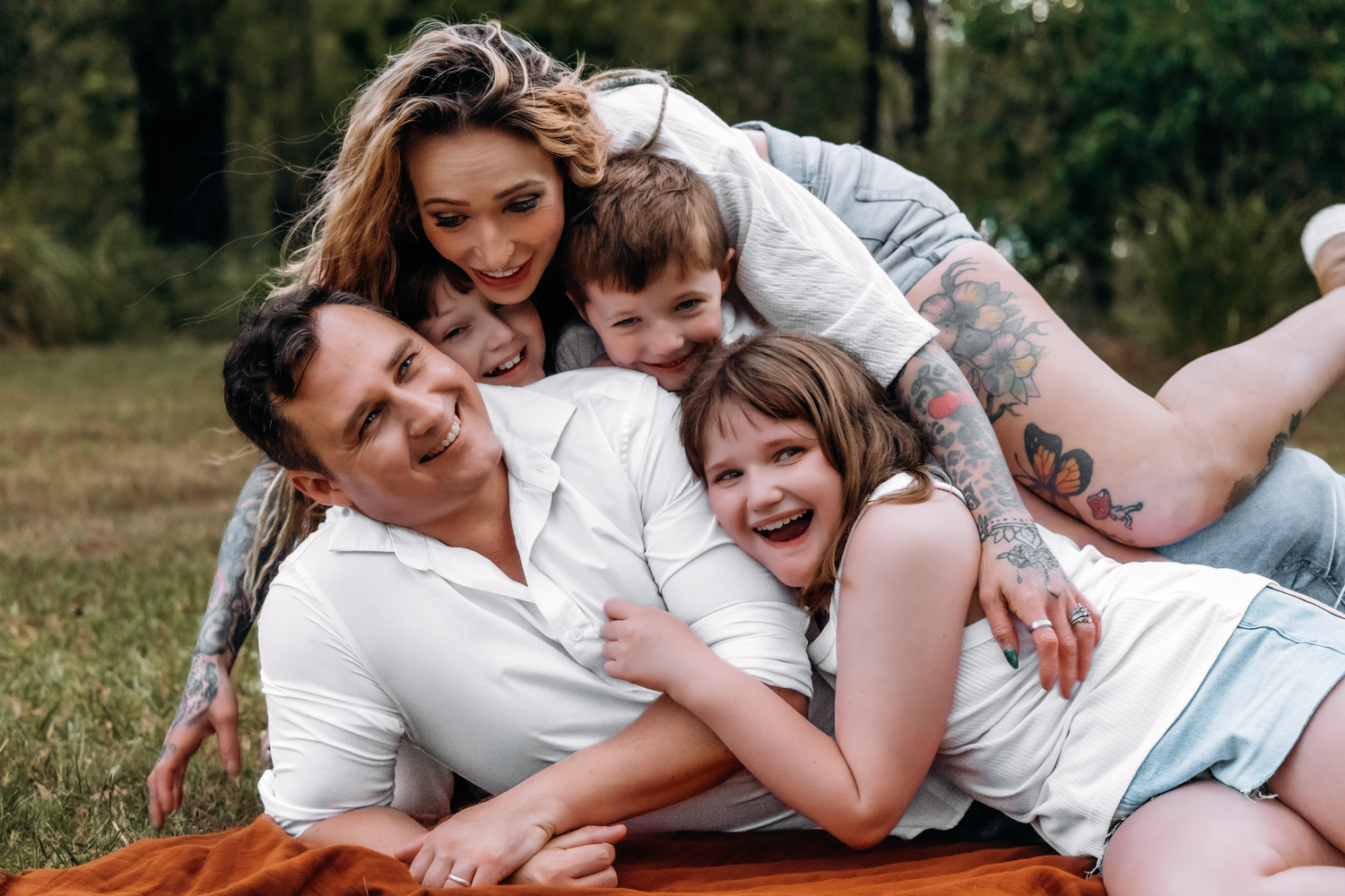 A tattooed couple laughs joyfully on a blanket in a park as two children pile on top of them.