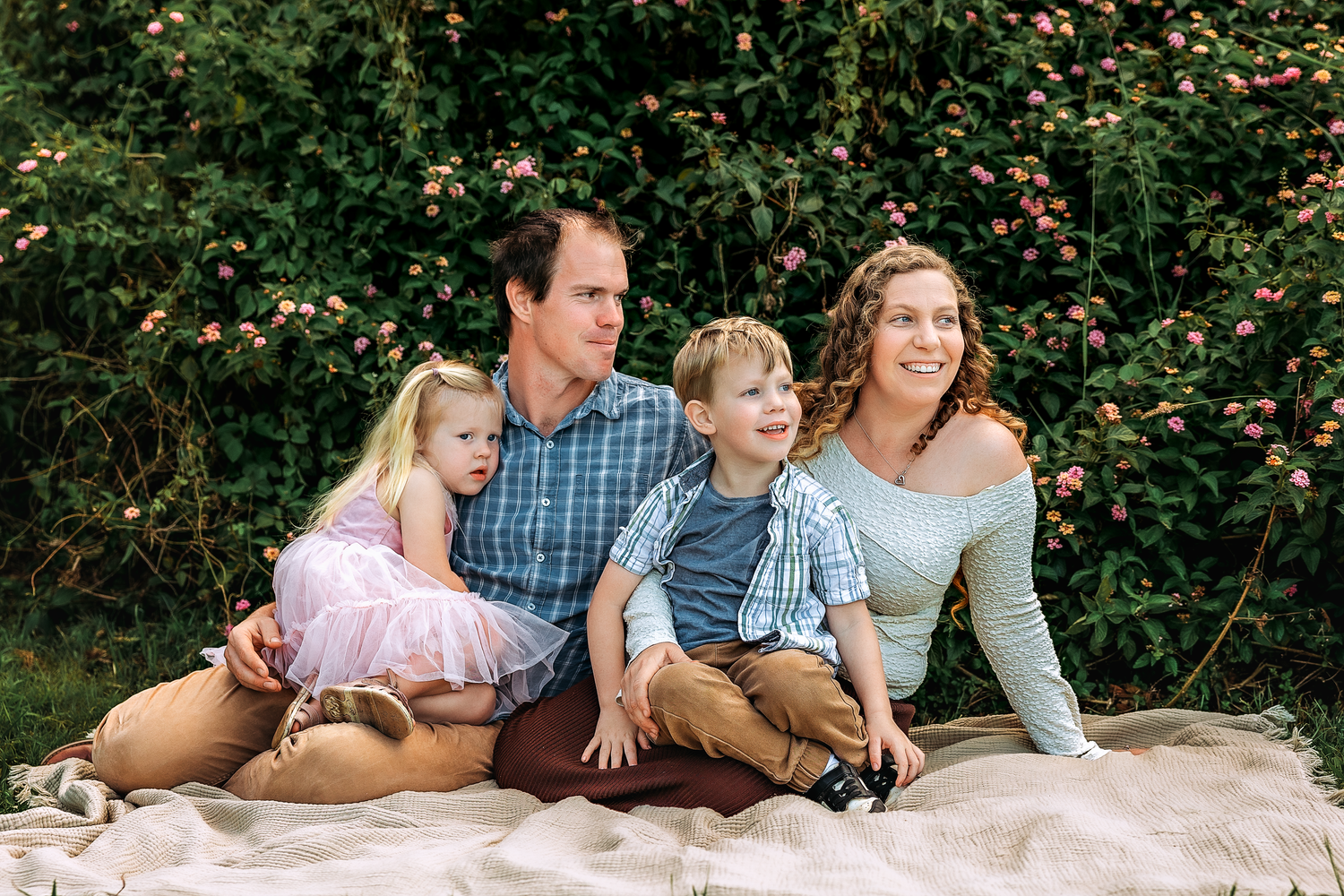 Happy family of four sitting on a blanket outdoors, smiling together near a lush green flowering bush.