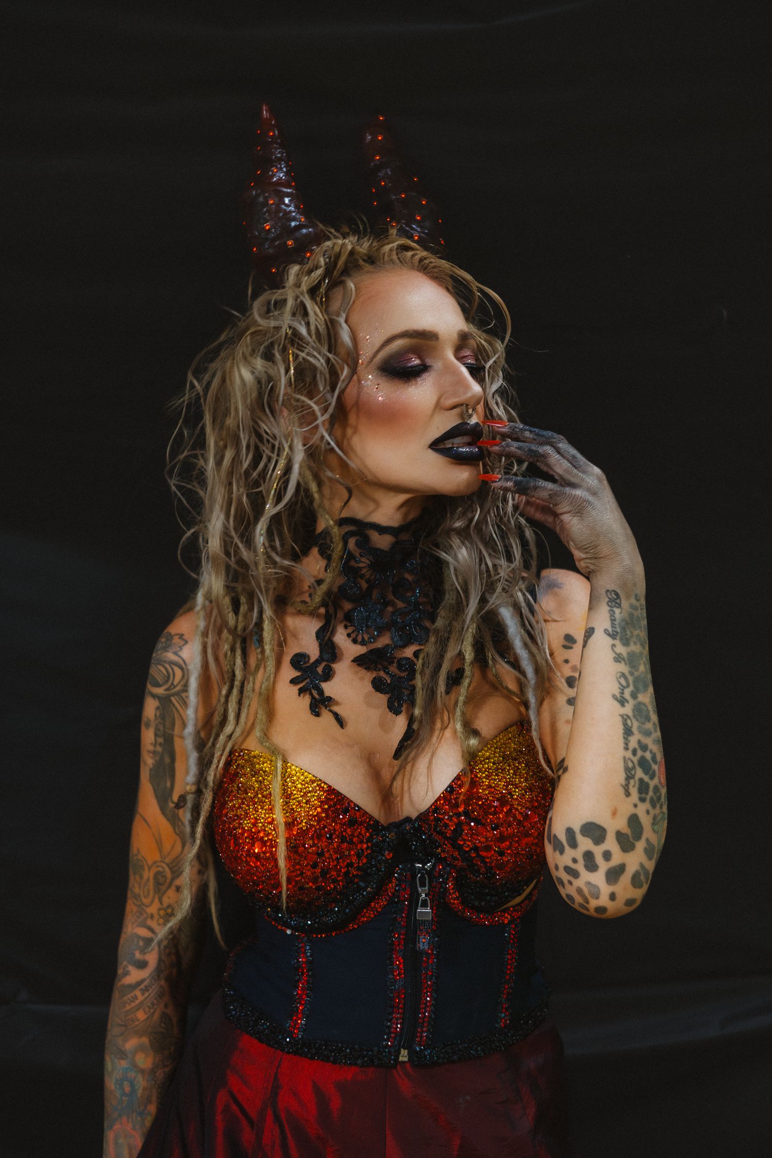 A tattooed woman in a red corset with devil horns and black lipstick poses on dark background.