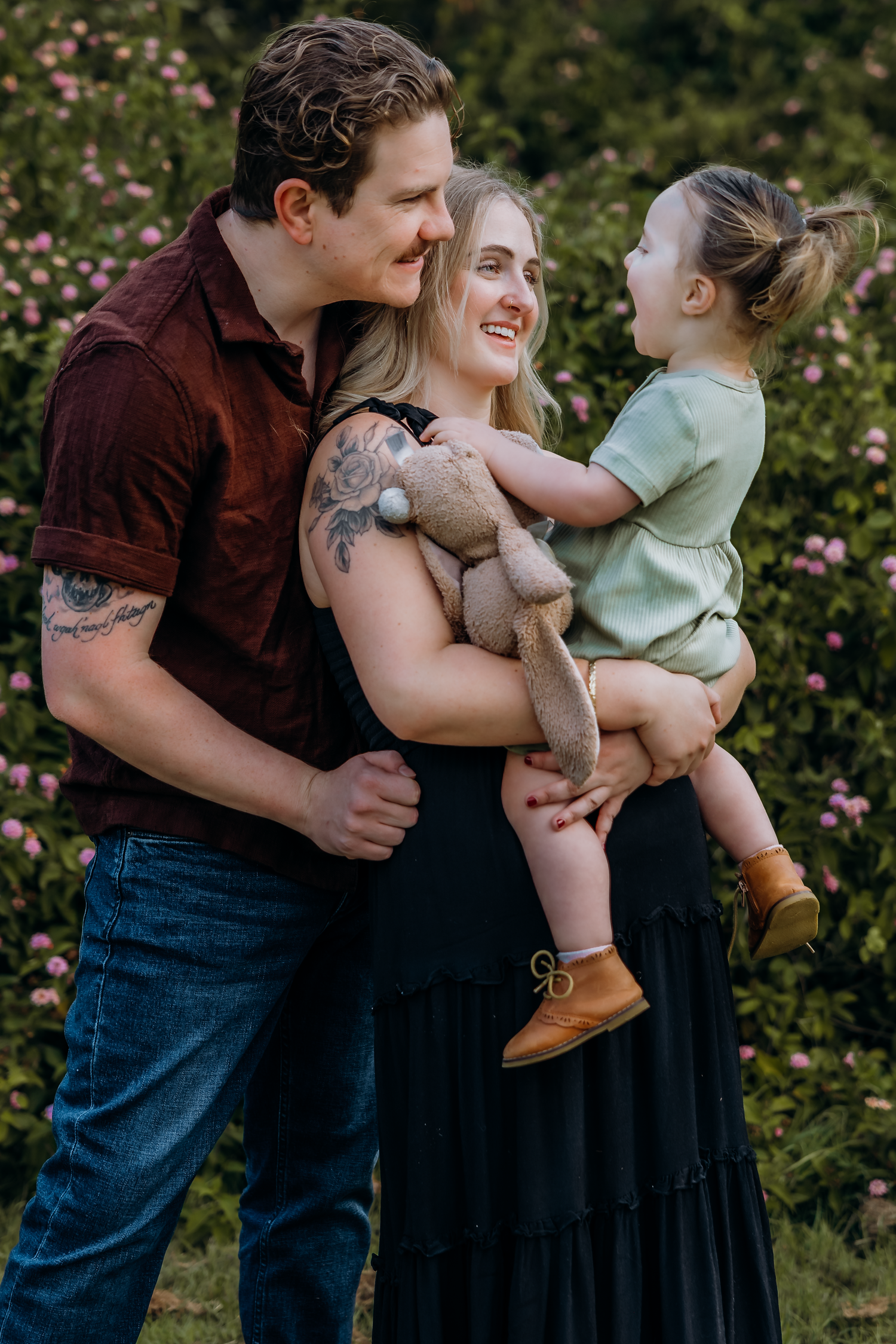 A happy young family with tattoos poses outdoors, mom holding toddler with stuffed bunny among pink blooms.