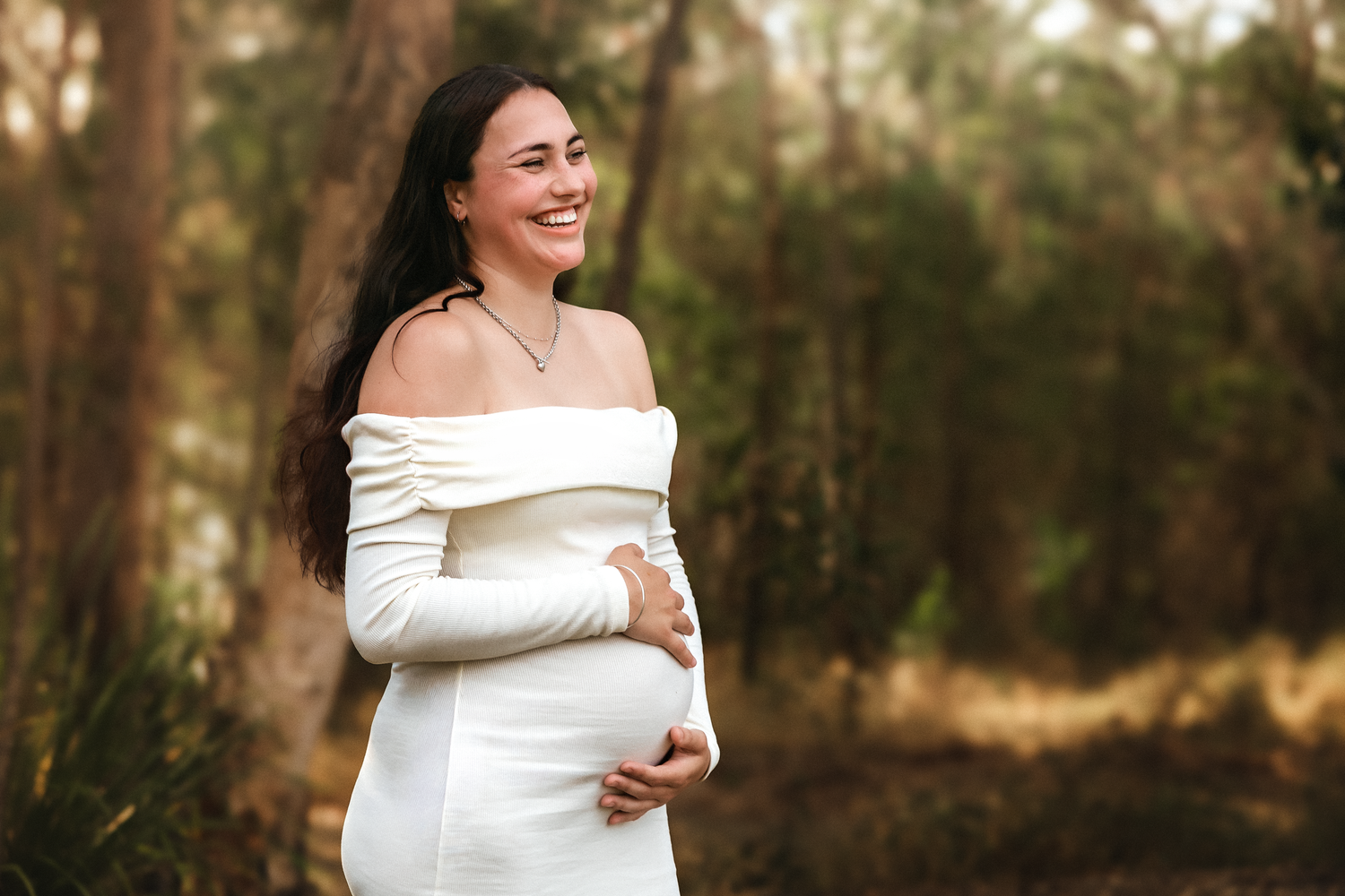 Smiling pregnant woman in white off-shoulder gown cradling baby bump outdoors in forest setting.