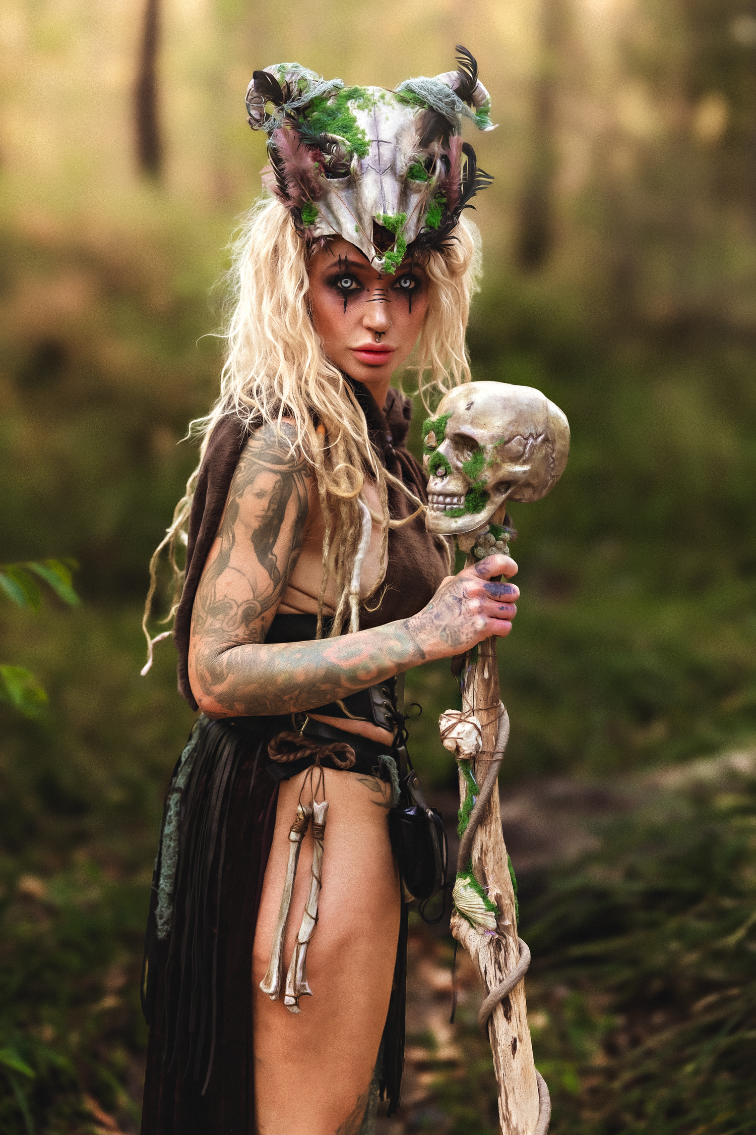 Tattooed blonde woman in fantasy forest warrior costume with animal skull headdress holding a staff topped with a human skull.