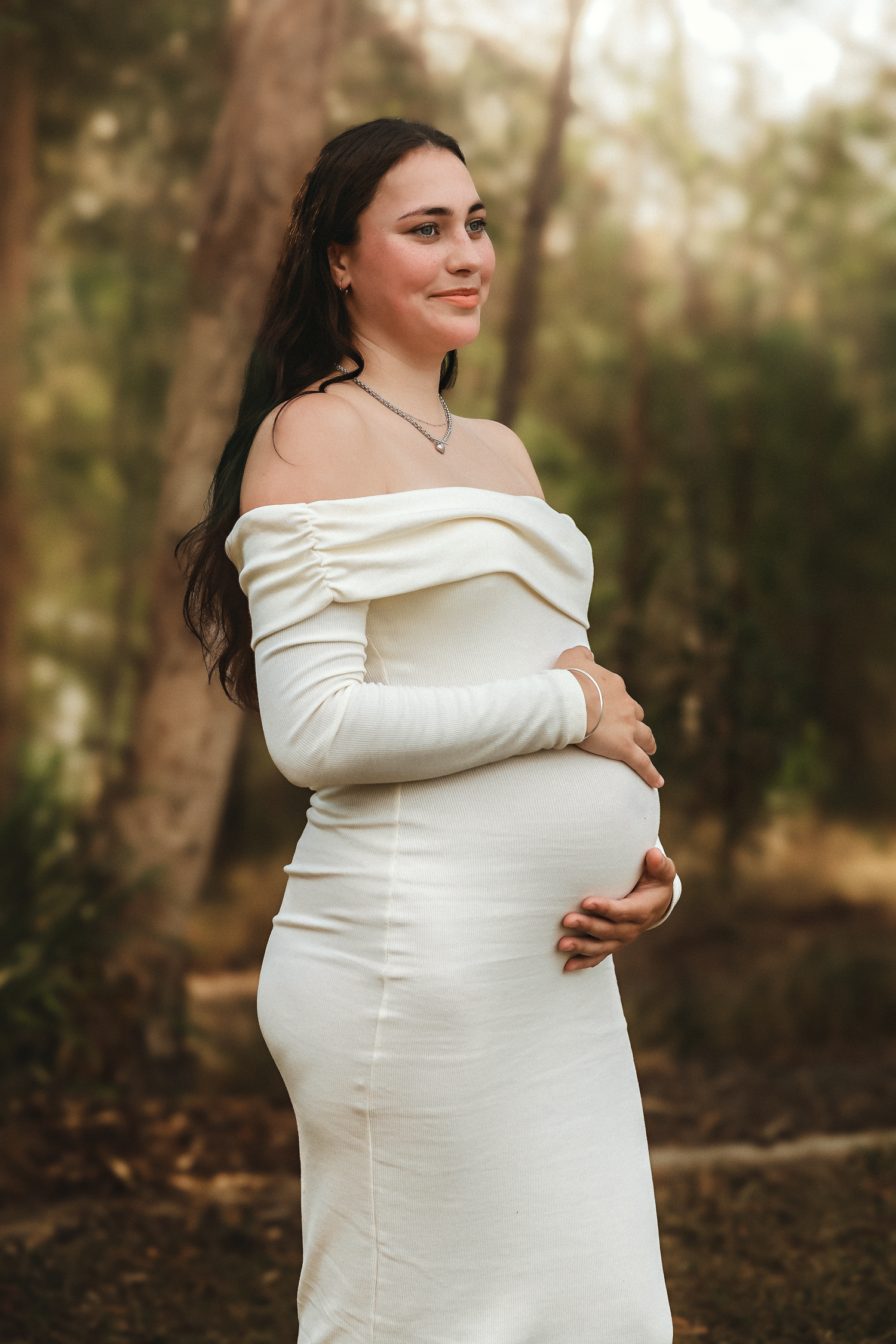 Pregnant woman in white off-shoulder gown cradling baby bump outdoors among trees during maternity photoshoot.