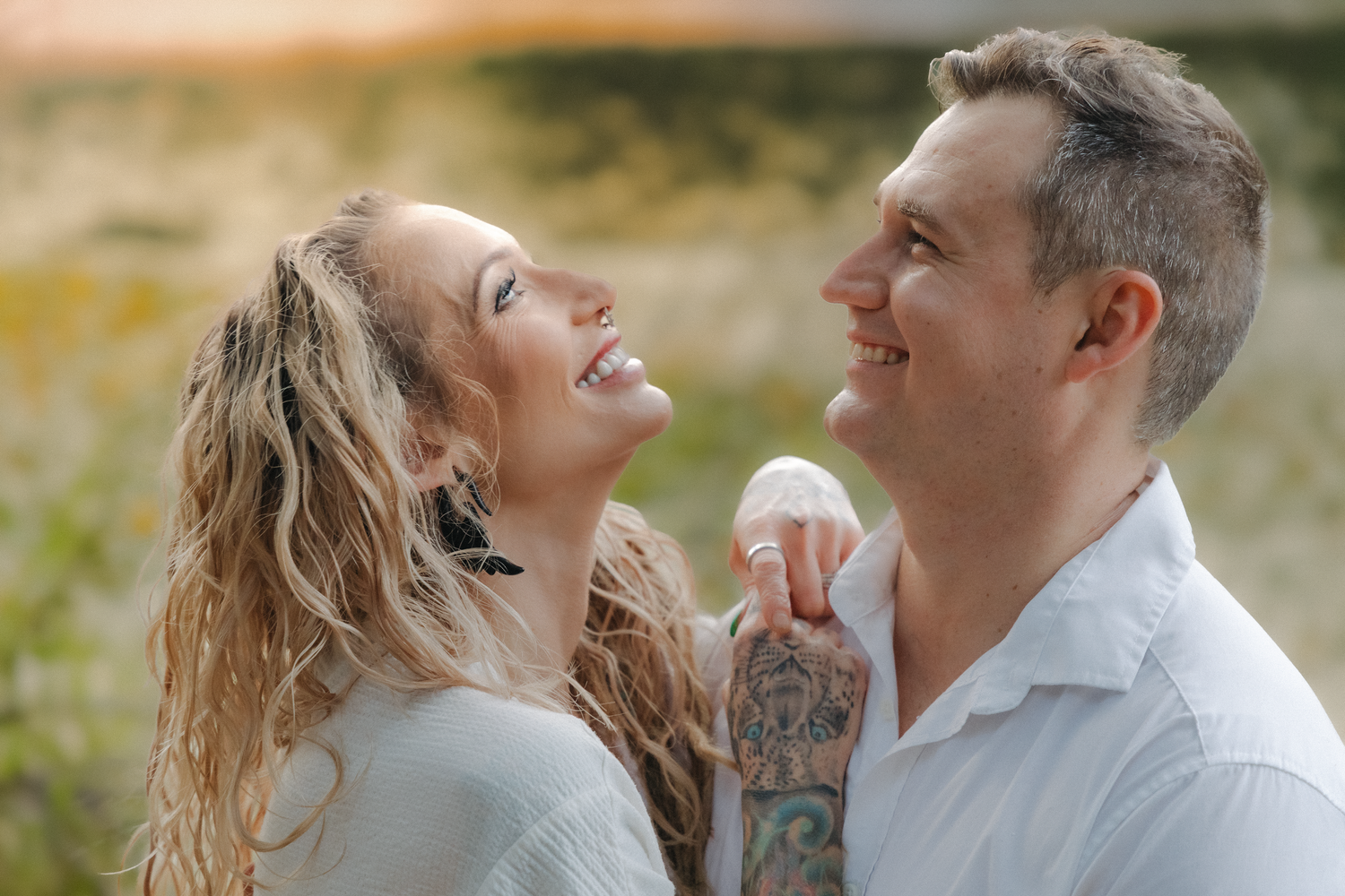 Smiling couple in golden hour light, woman with curly blonde hair and man in white shirt with tattoed arm embracing outdoors.