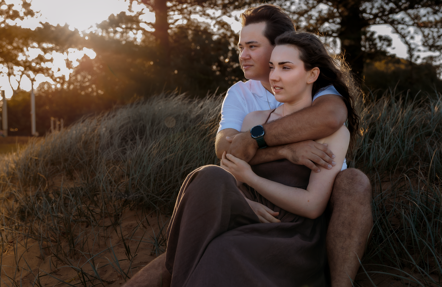 Couple embracing at sunset on sandy dunes, golden light filtering through trees in a romantic outdoor setting.