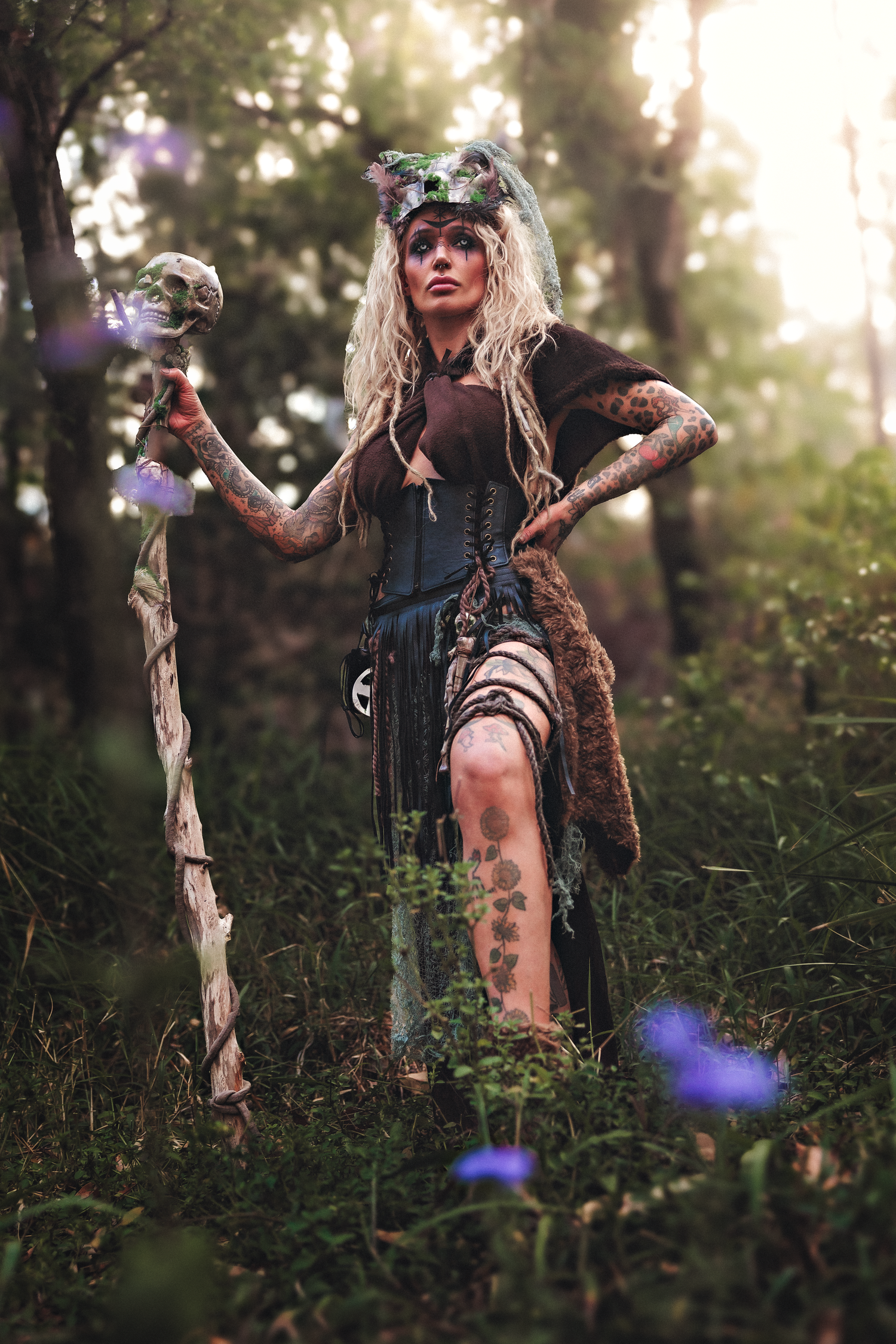 A tattooed woman in fantasy forest witch costume with skull staff and floral headdress poses in a mystical woodland setting.