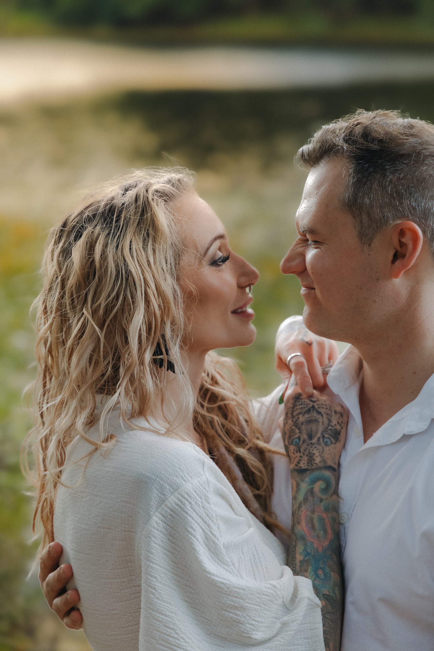 Couple sharing a romantic moment near a lake, woman with blonde wavy hair in white dress, man with tattoed arm.