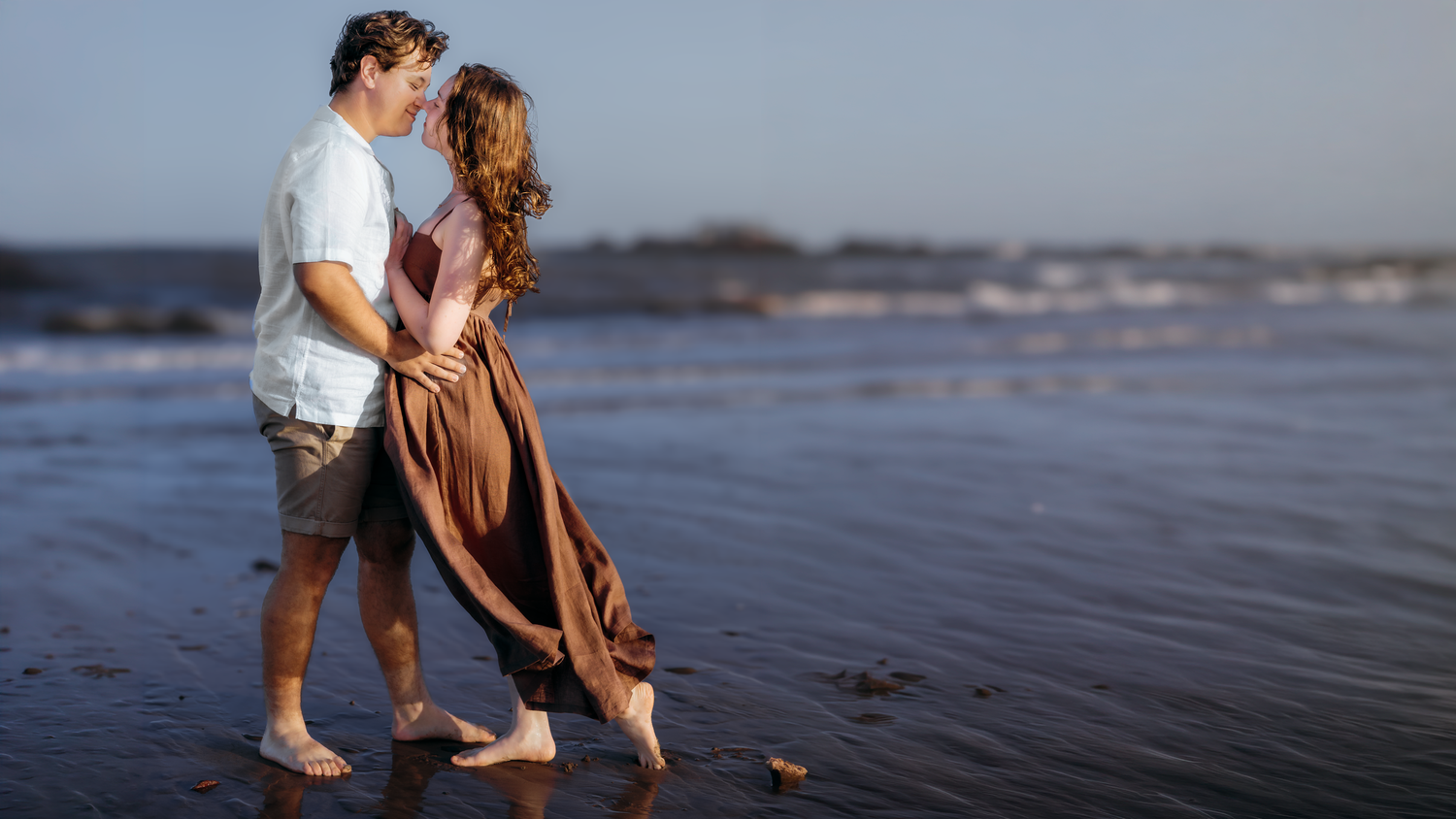 A romantic couple embraces barefoot on a serene beach at sunset, woman in flowing brown dress.