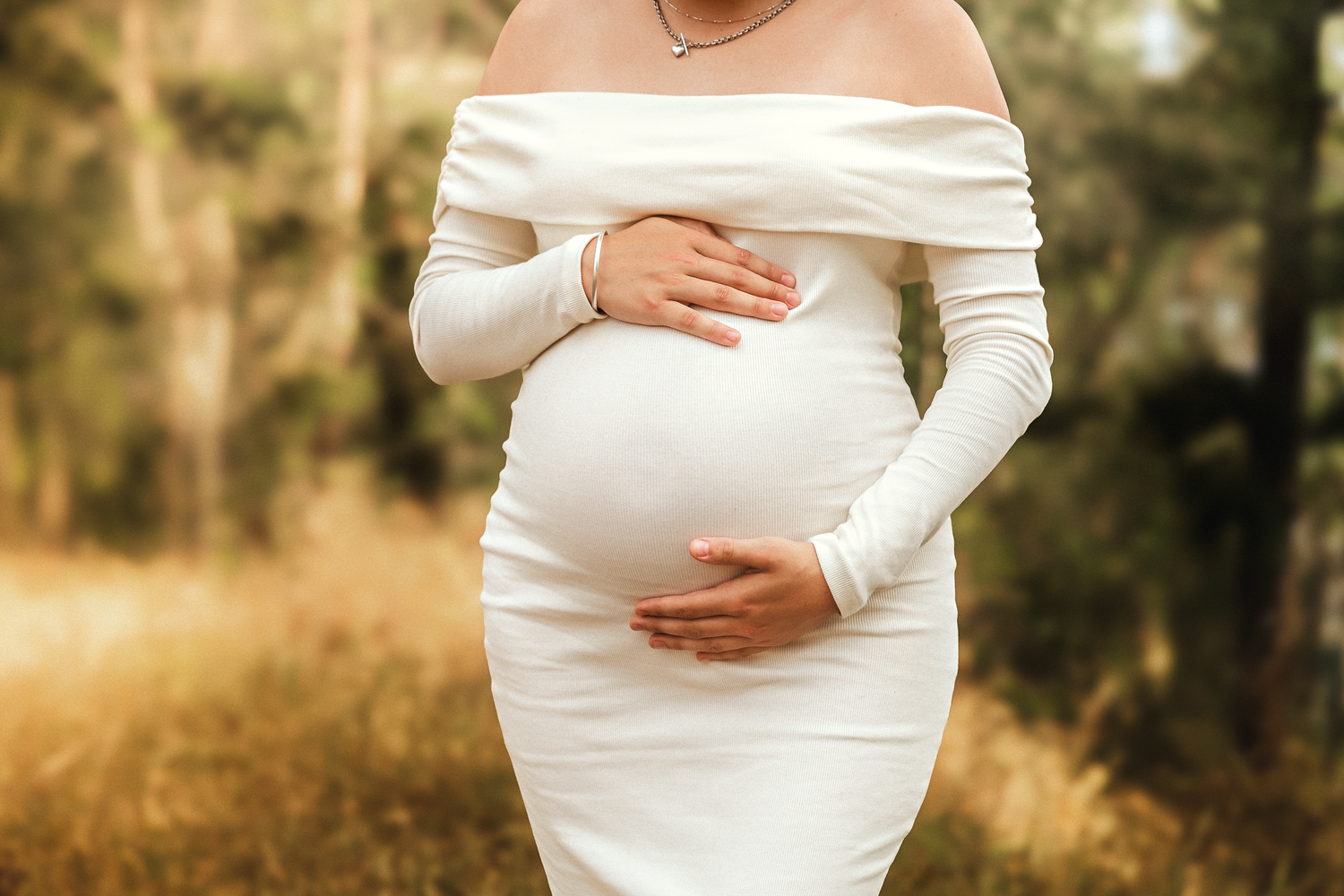 Pregnant woman in white off-shoulder dress cradling baby bump outdoors in golden natural light.
