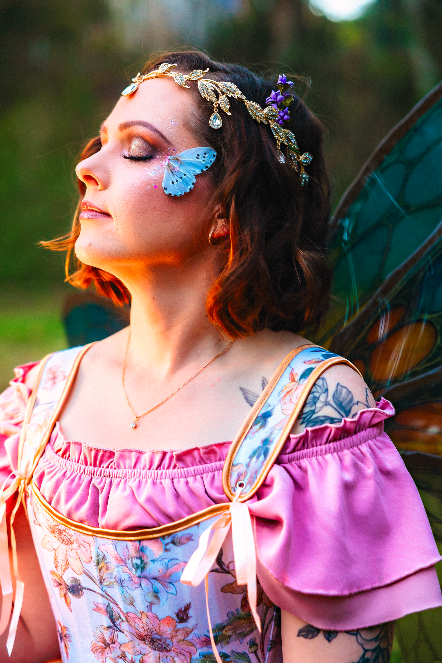 A woman dressed as a fairy with butterfly face paint, floral crown, pink dress, and teal wings in a forest setting.