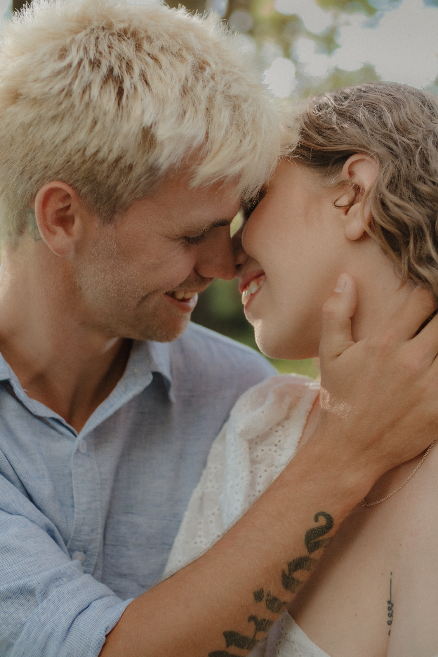 A couple smiles forehead-to-forehead outdoors, the man with a tattoo gently cupping her face.