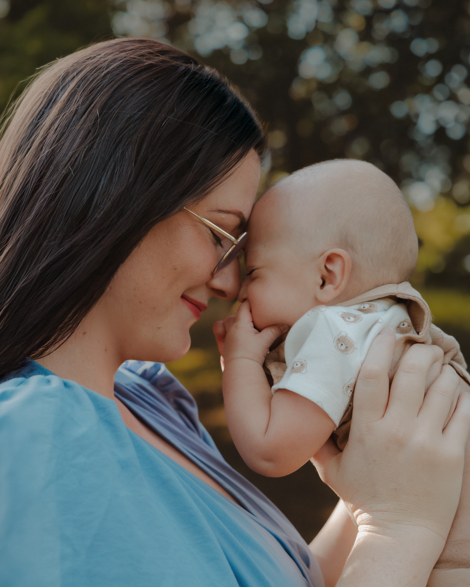 A dark-haired mother with glasses gently presses her forehead against her baby in warm sunlight.