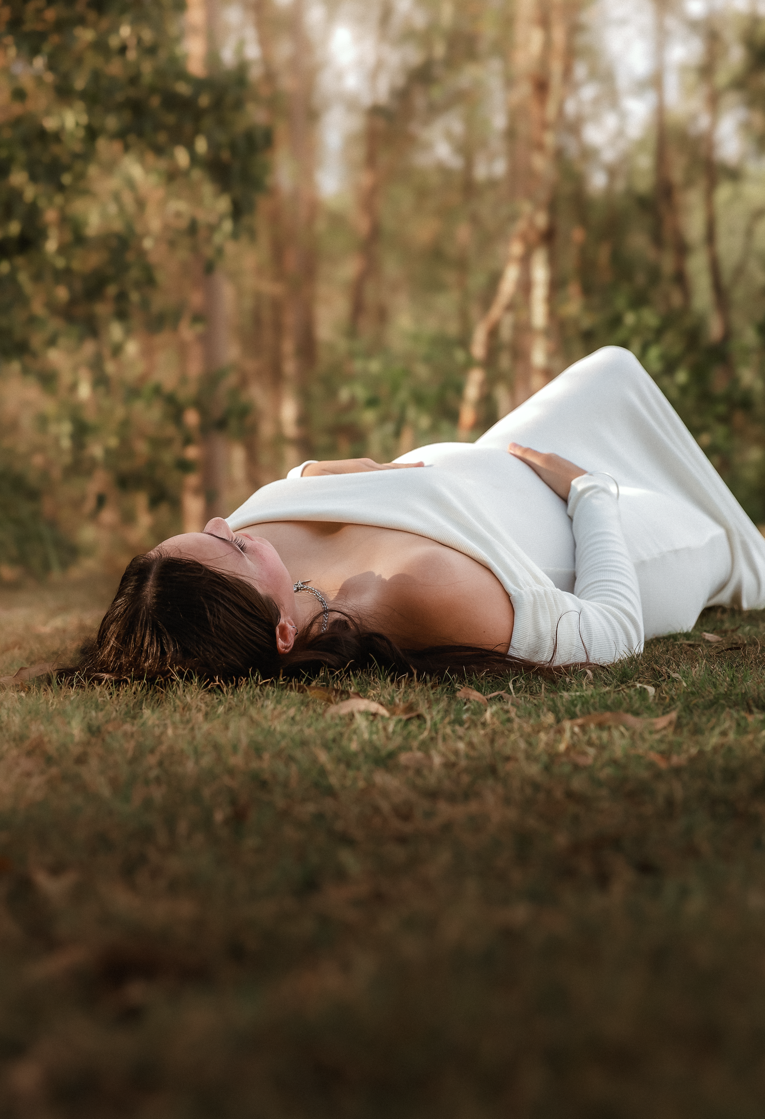 A woman in a white dress lies on green grass in a serene forest setting, gazing upward peacefully.