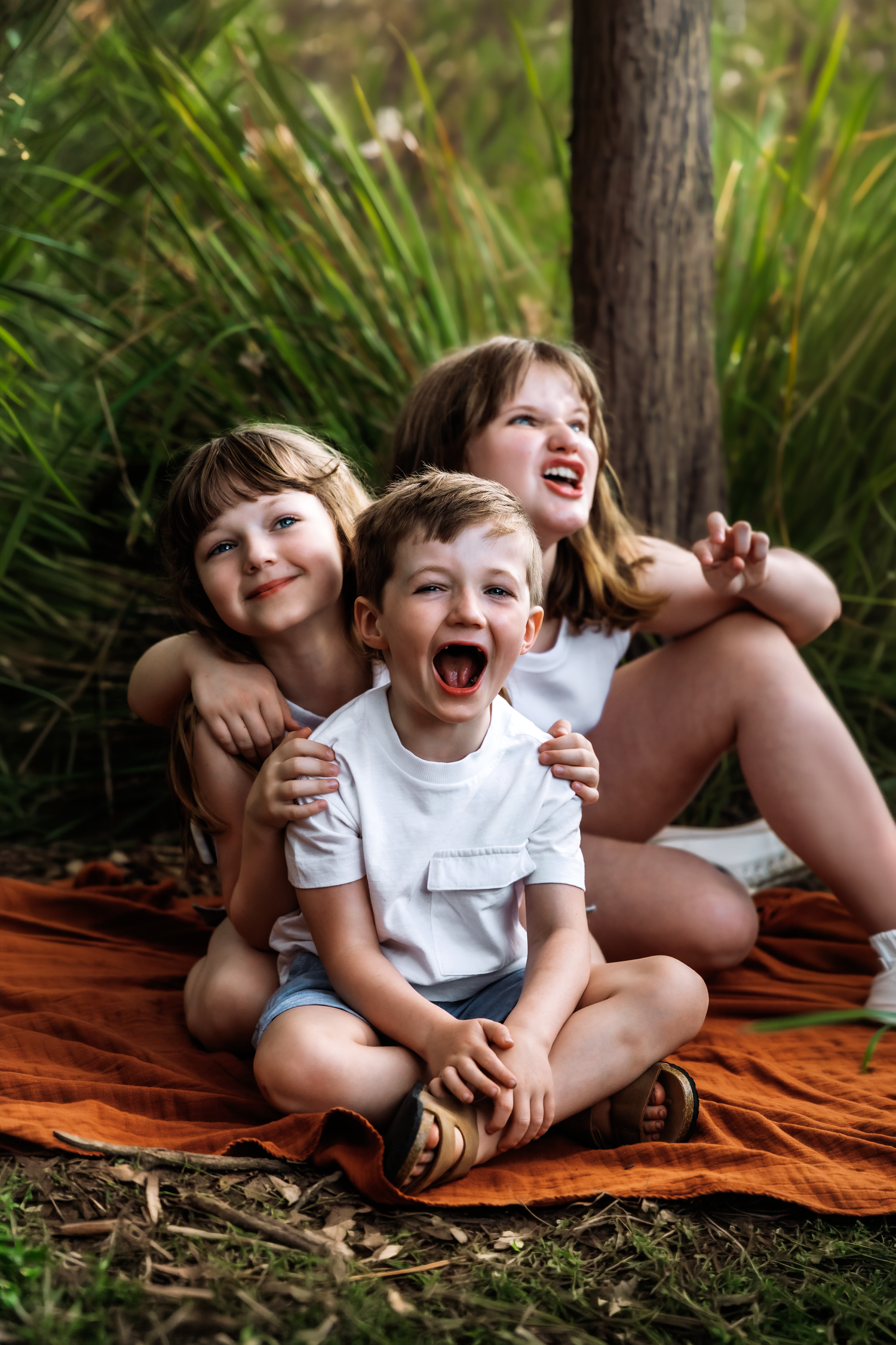 Three happy children sitting on an orange blanket outdoors, laughing and embracing near green plants and a tree.
