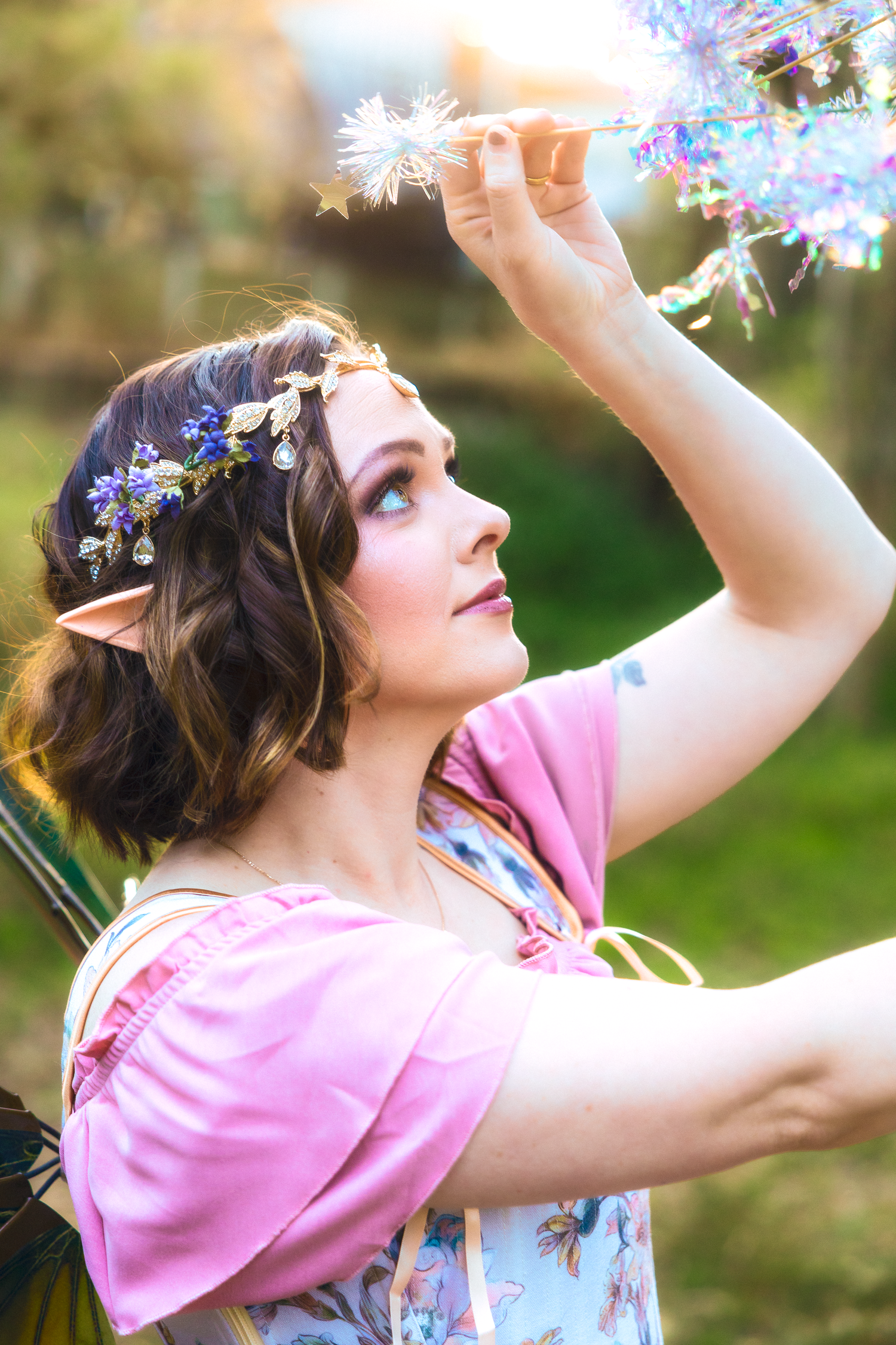 A woman in a pink dress with elf ears and floral crown reaches up to touch sparkling decorations outdoors.