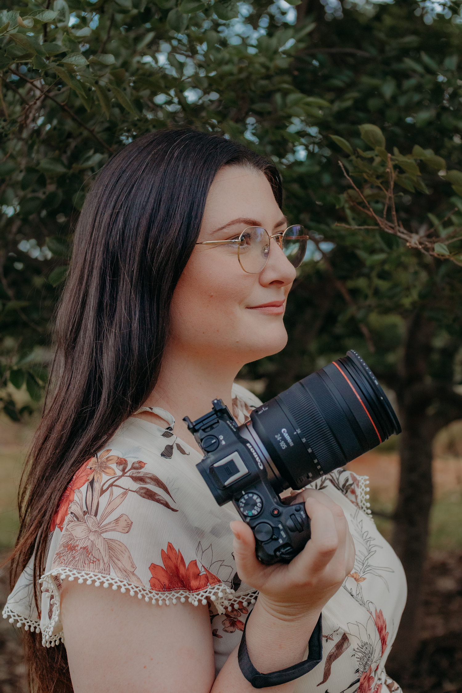 A dark-haired woman with glasses holds a Canon camera outdoors among green trees.
