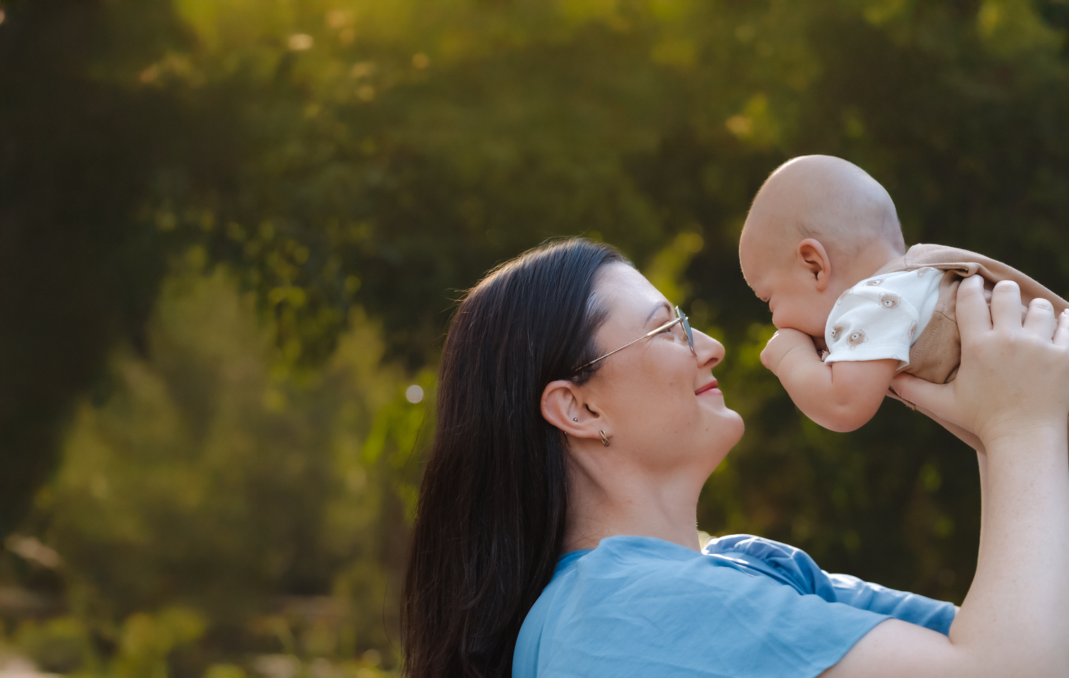 A mother joyfully lifts her baby in the air outdoors, sharing a tender moment surrounded by lush green trees.