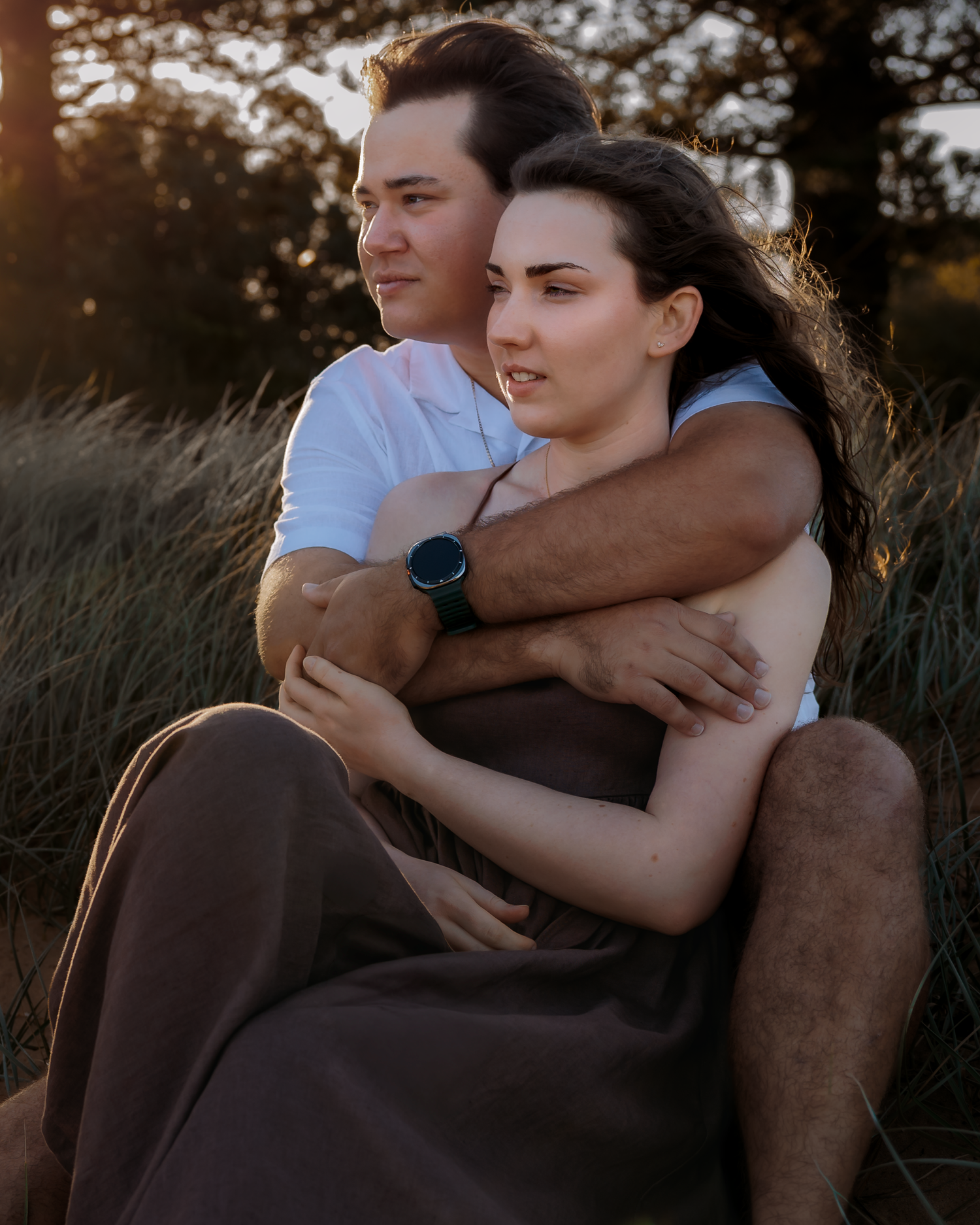 A couple embraces at golden hour in a grassy field, the man wrapping his arms around the woman from behind.