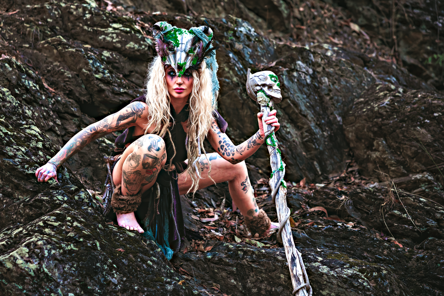 Tattooed woman in fantasy wolf headdress and tribal costume poses dramatically on rocky terrain holding a decorated staff.