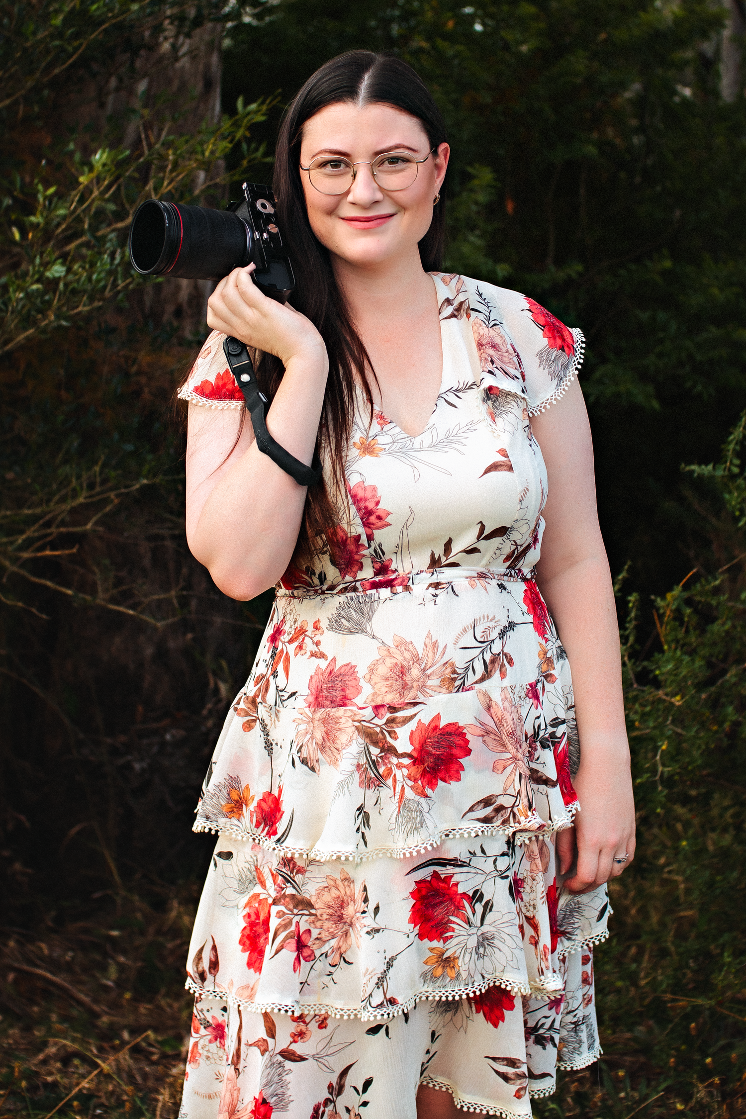Woman in floral dress holding a camera outdoors, smiling, wearing glasses, surrounded by lush green foliage.