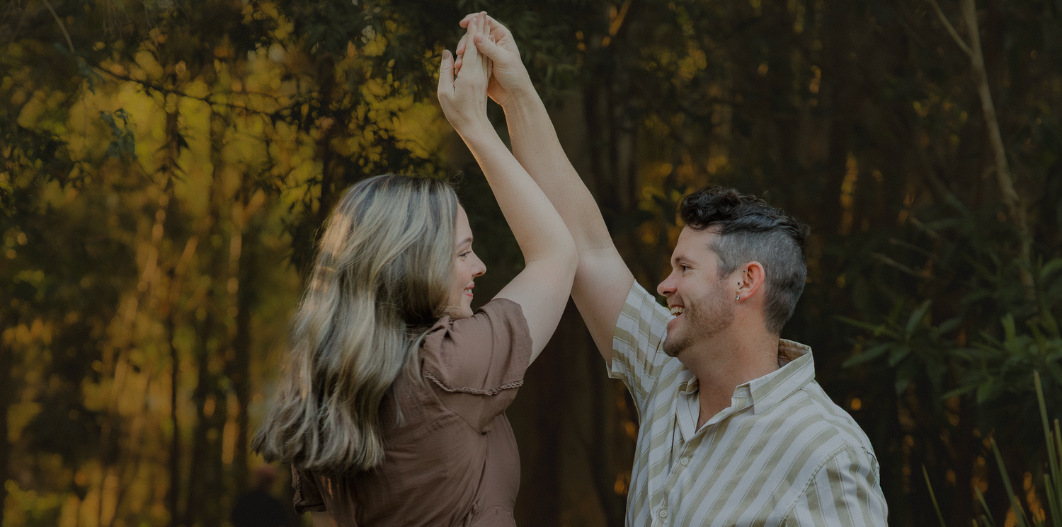 Joyful couple dancing and spinning hands in a sunlit forest, laughing together in casual autumn outfits.