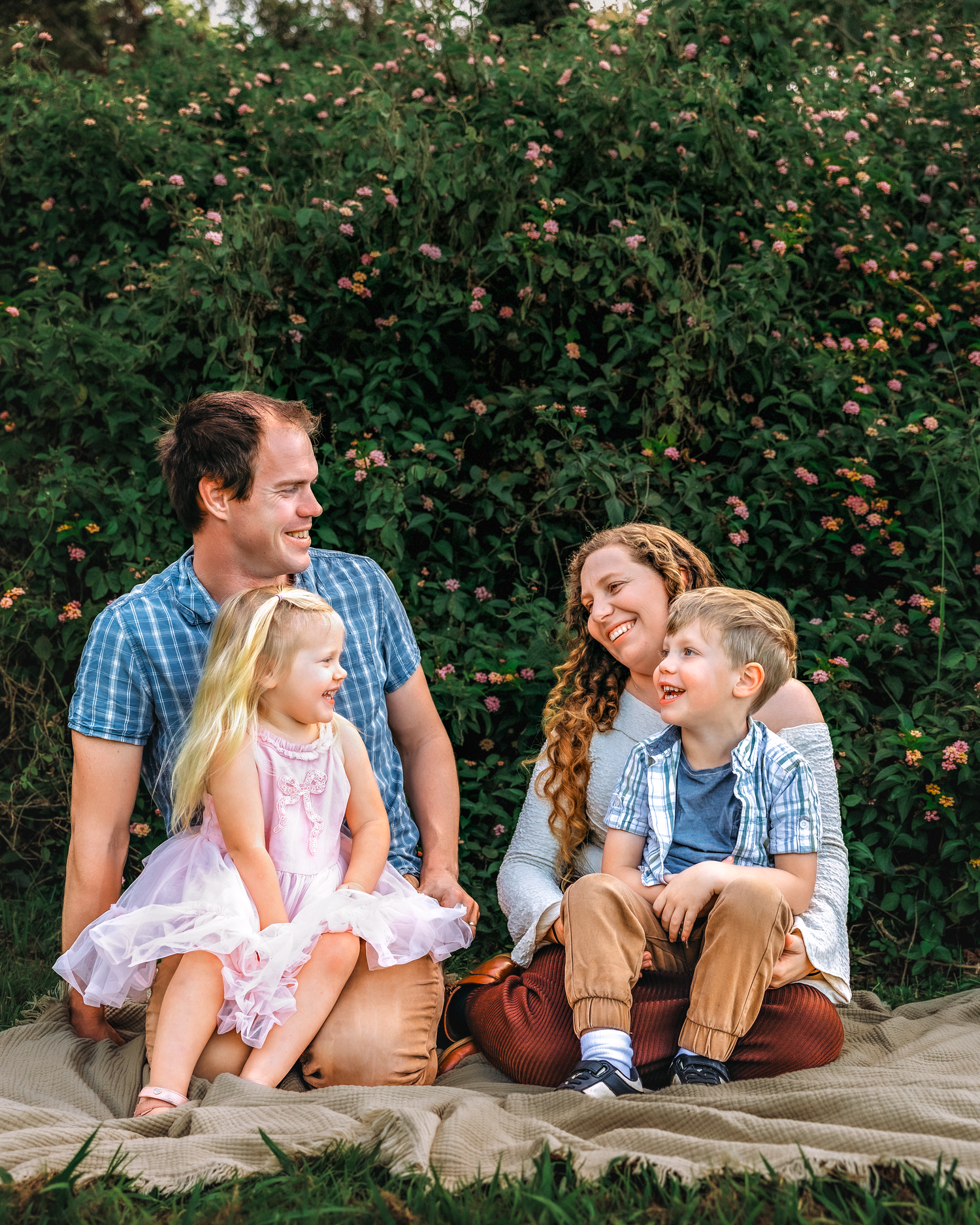 Happy family of four sitting outdoors on a blanket, smiling together in front of a lush green flowering hedge.