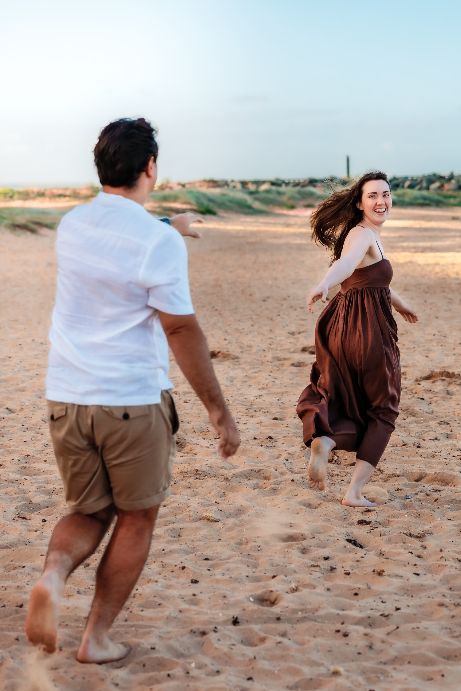 A joyful couple runs barefoot on sandy beach, woman in brown dress laughing as man chases her at sunset.