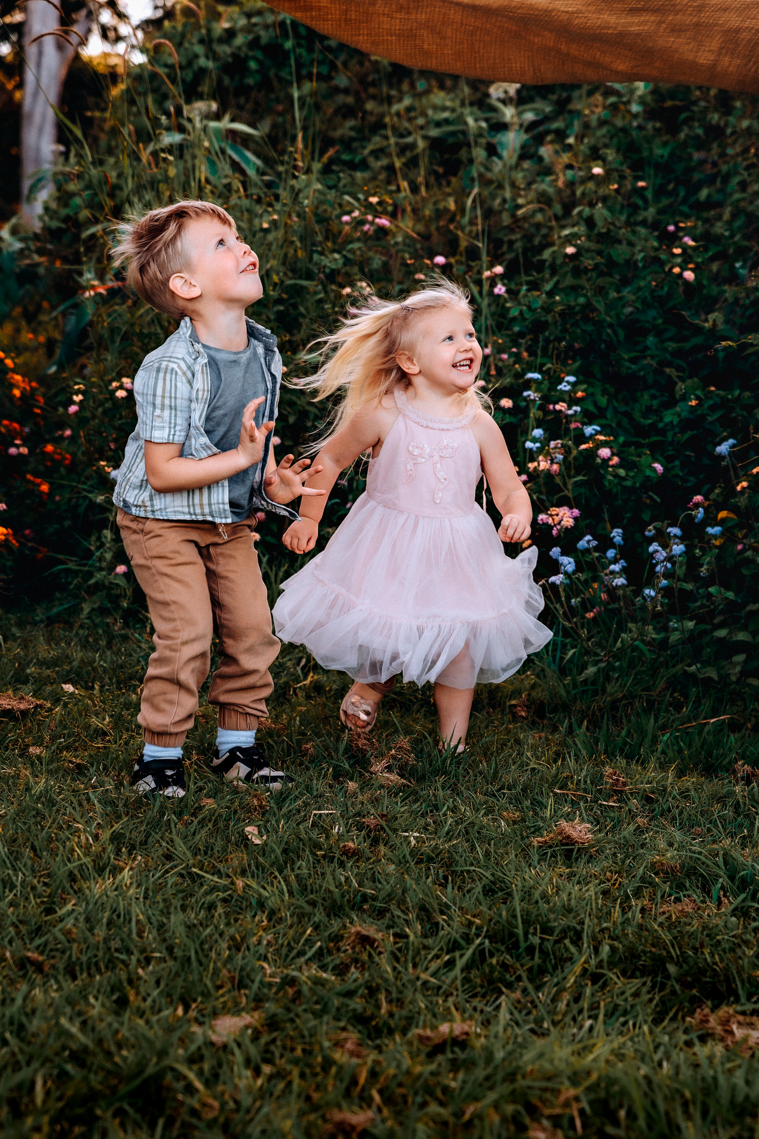 Two young children laughing and playing outdoors in a garden, boy in plaid shirt and girl in pink tulle dress.