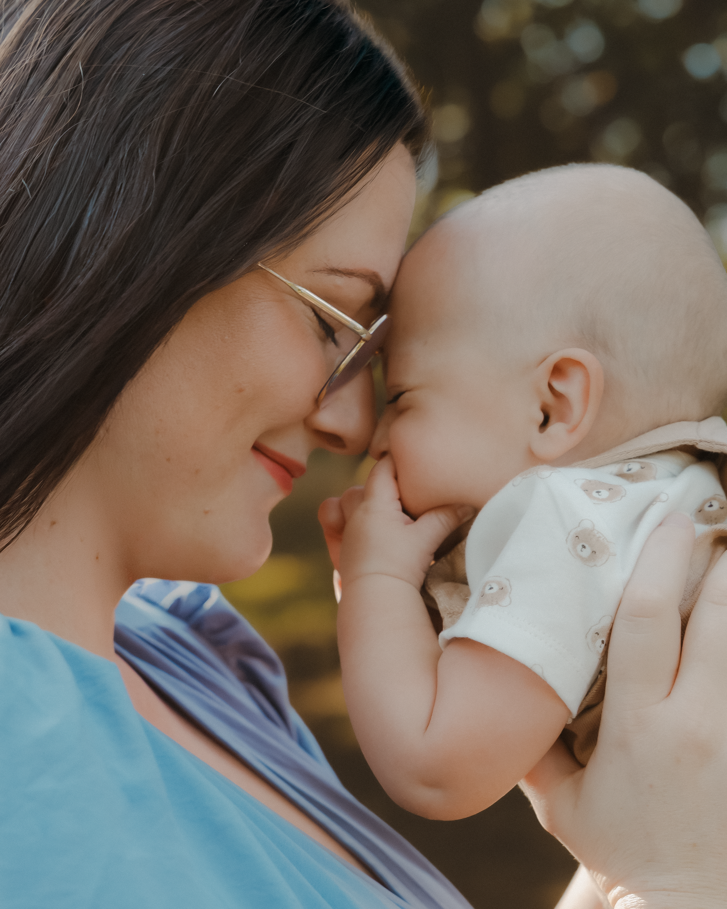A mother tenderly touches foreheads with her smiling baby outdoors in warm sunlight.