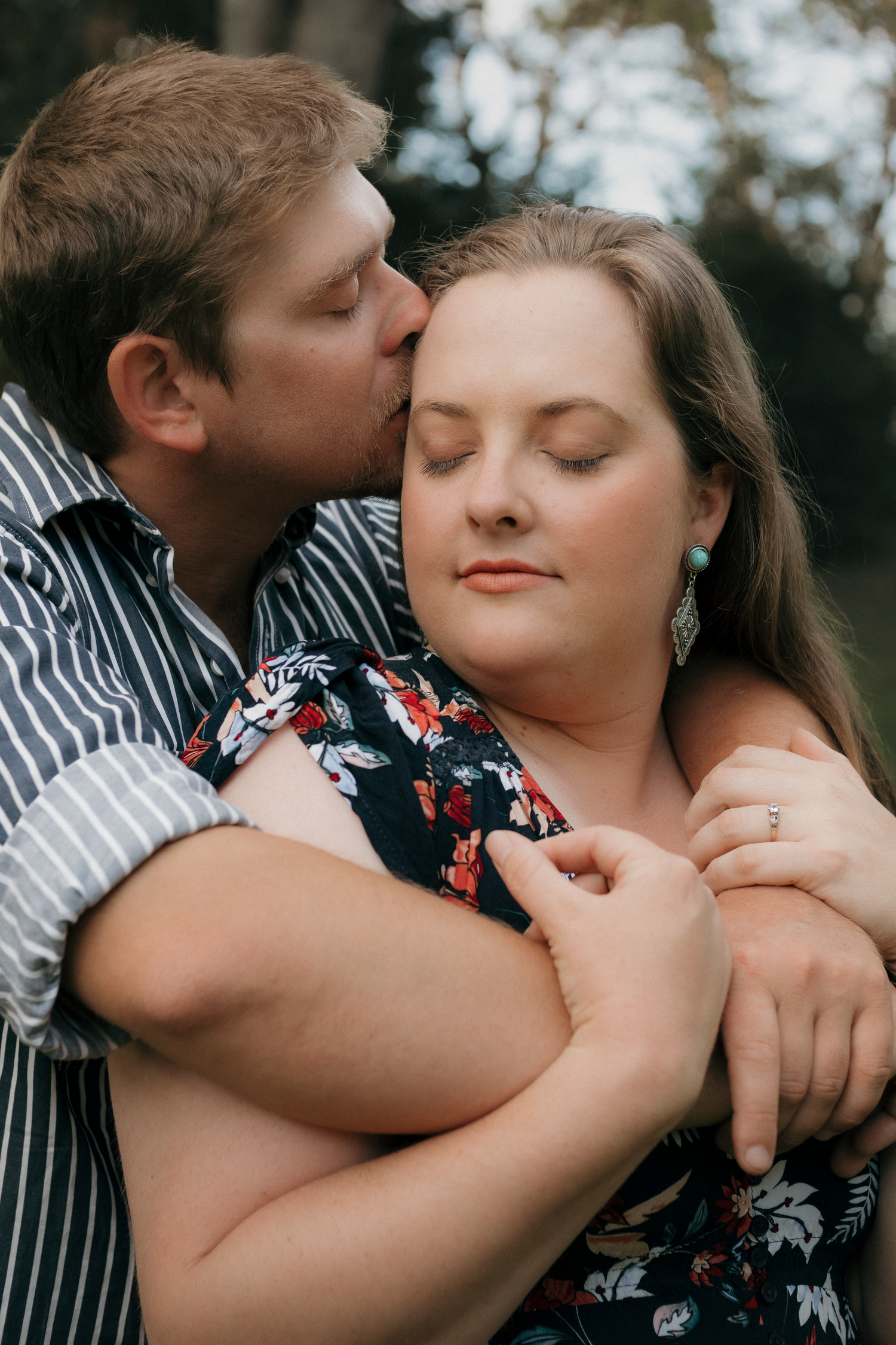 Couple in outdoor setting, man kissing tattooed woman on cheek while embracing her.