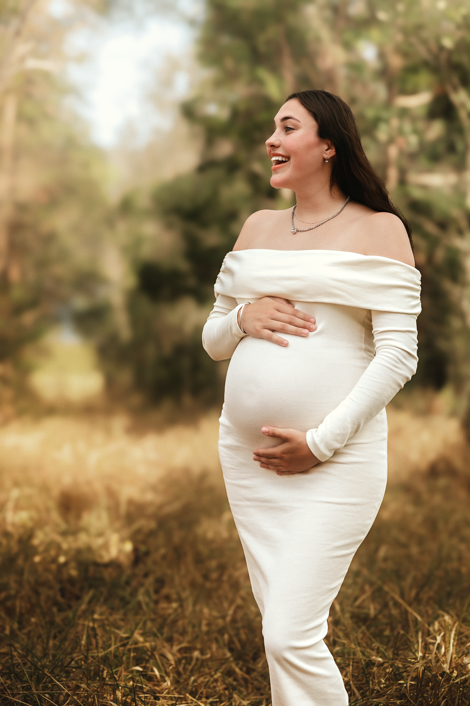 Smiling pregnant woman in white off-shoulder gown cradling her baby bump outdoors in a golden forest setting.