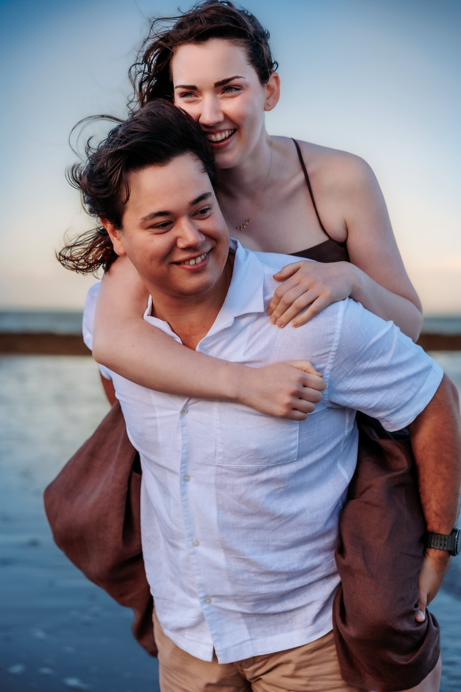 Happy couple at beach sunset, woman piggyback riding on mans back, both smiling joyfully near ocean.