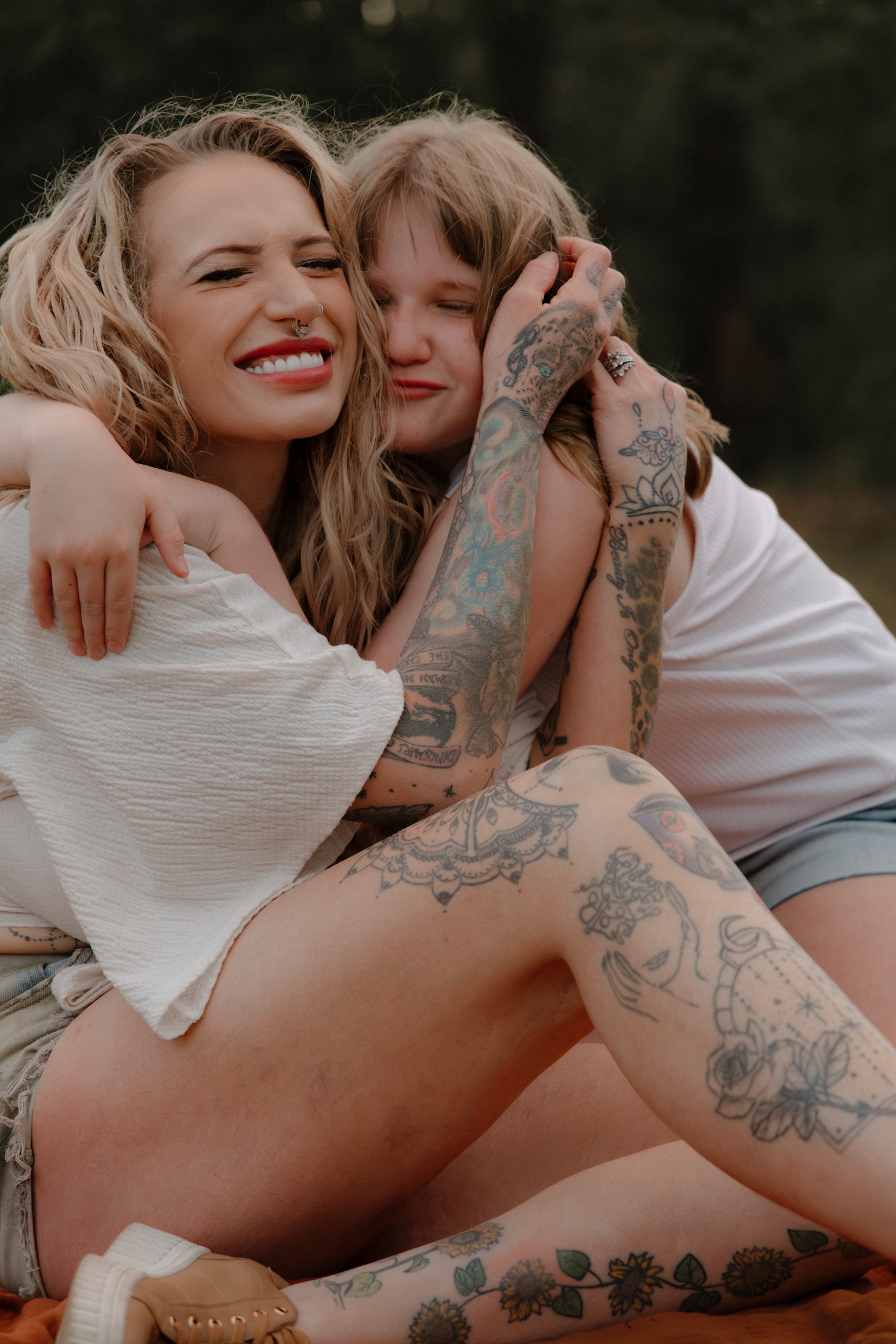 Two tattooed women embracing and smiling warmly while sitting outdoors in casual summer clothing among nature.
