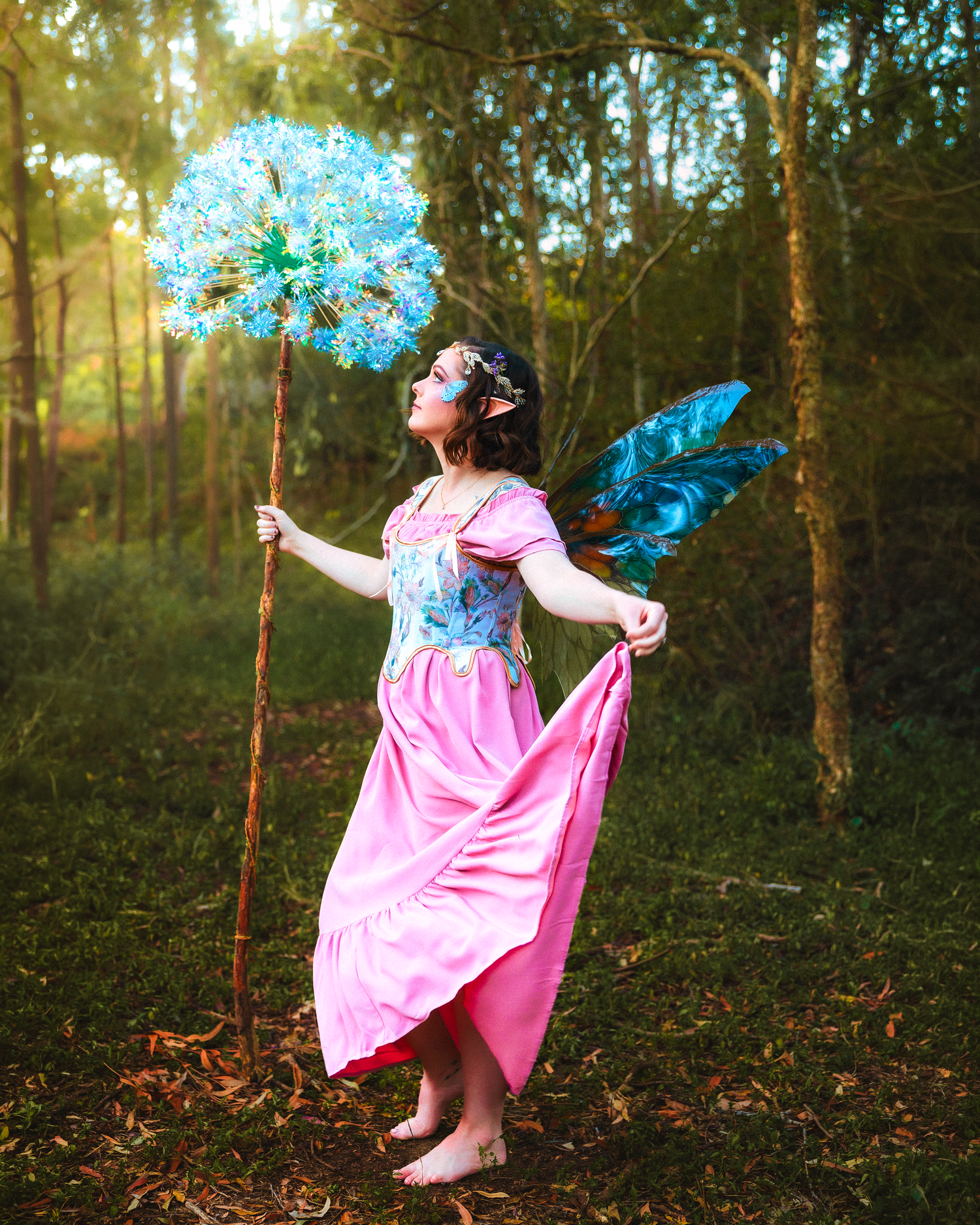 A girl in a pink fairy dress with blue wings holds a flower staff in an enchanted forest setting.