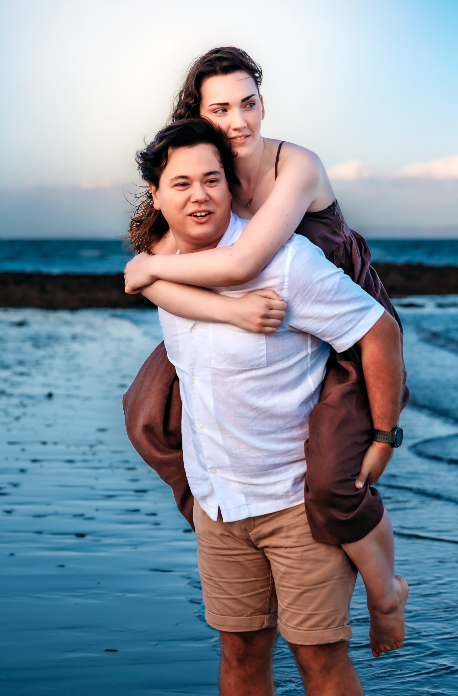 Couple posing on a beach at sunset, man giving woman a piggyback ride, both smiling, wearing casual summer clothes.