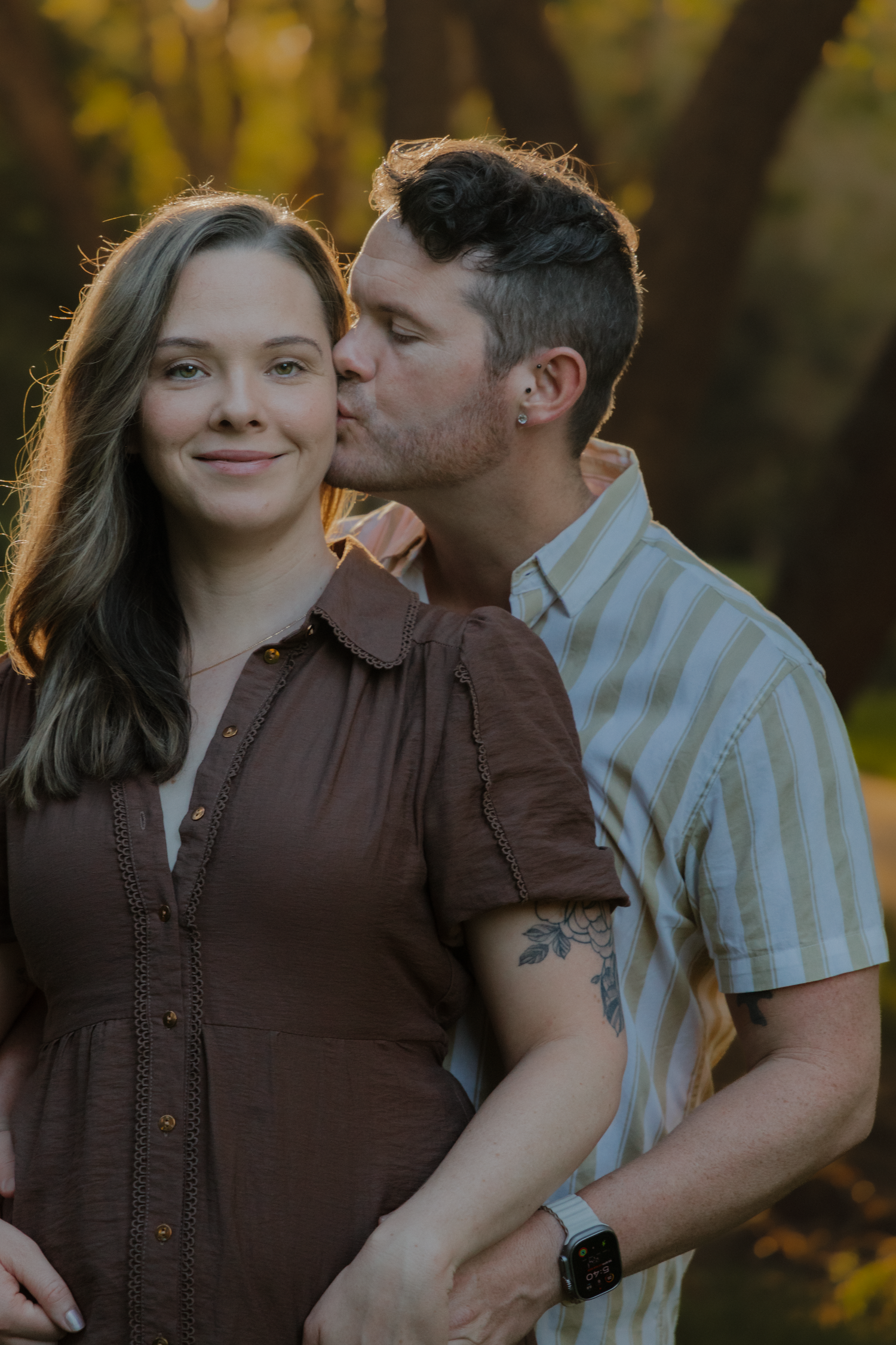 Man in striped shirt kissing woman on cheek outdoors during golden hour, woman smiling softly in brown dress.