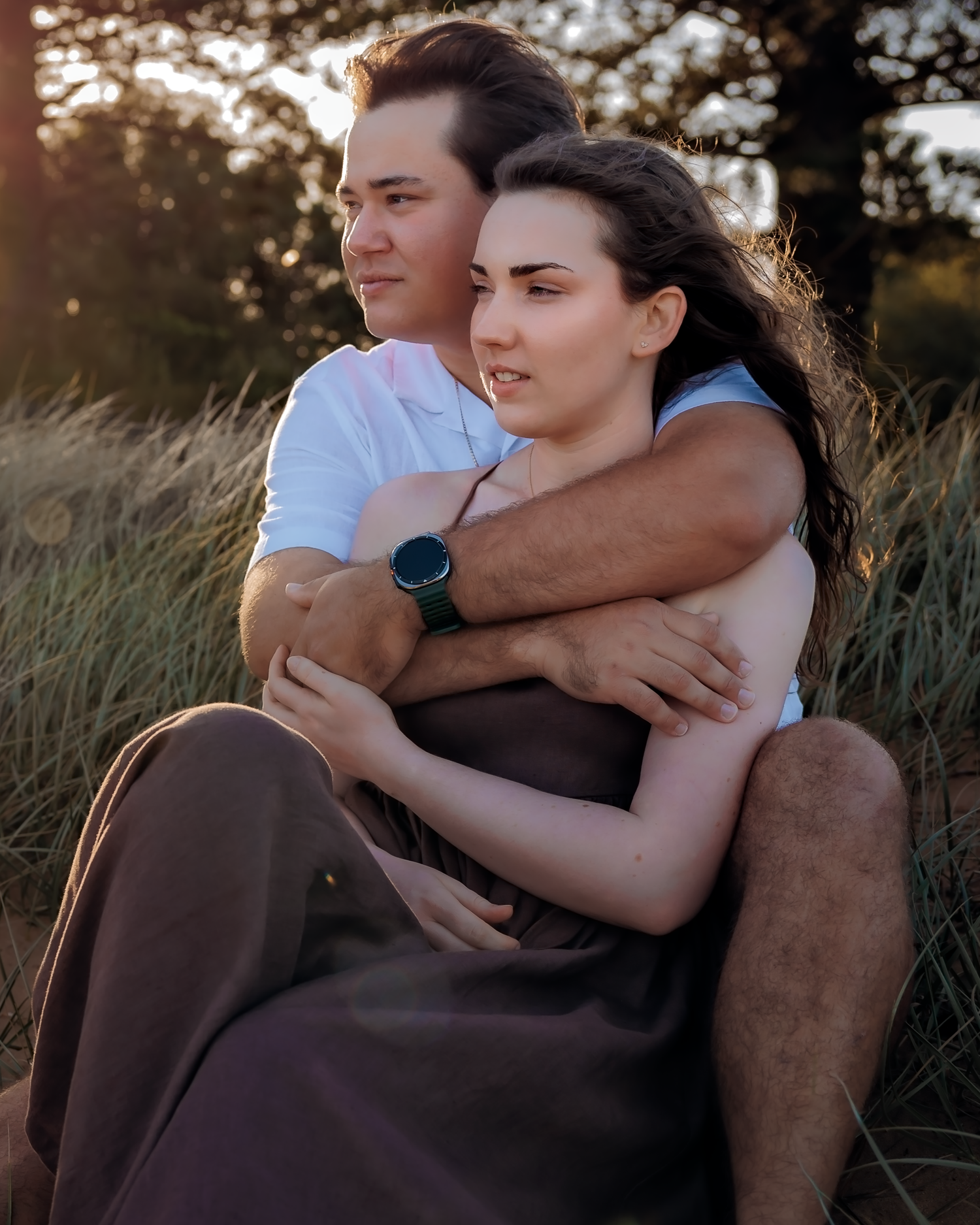 A couple sits together in a golden field at sunset, the man embracing the woman from behind as she gazes into the distance.