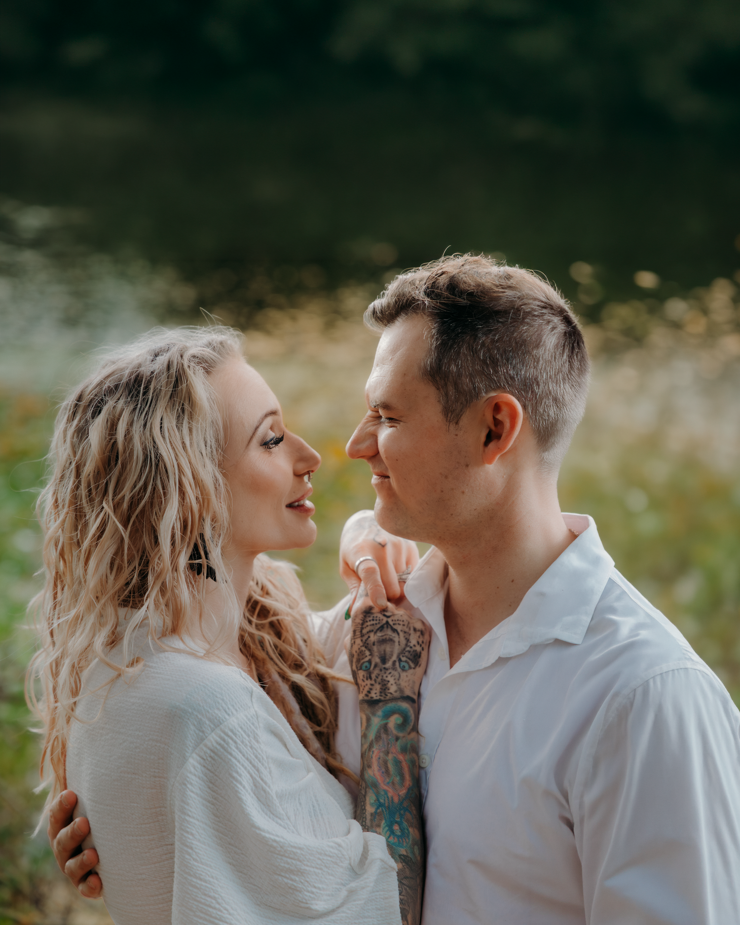 Romantic couple gazing at each other near a lake, man with tattoed arm in white shirt, woman with blonde curls.