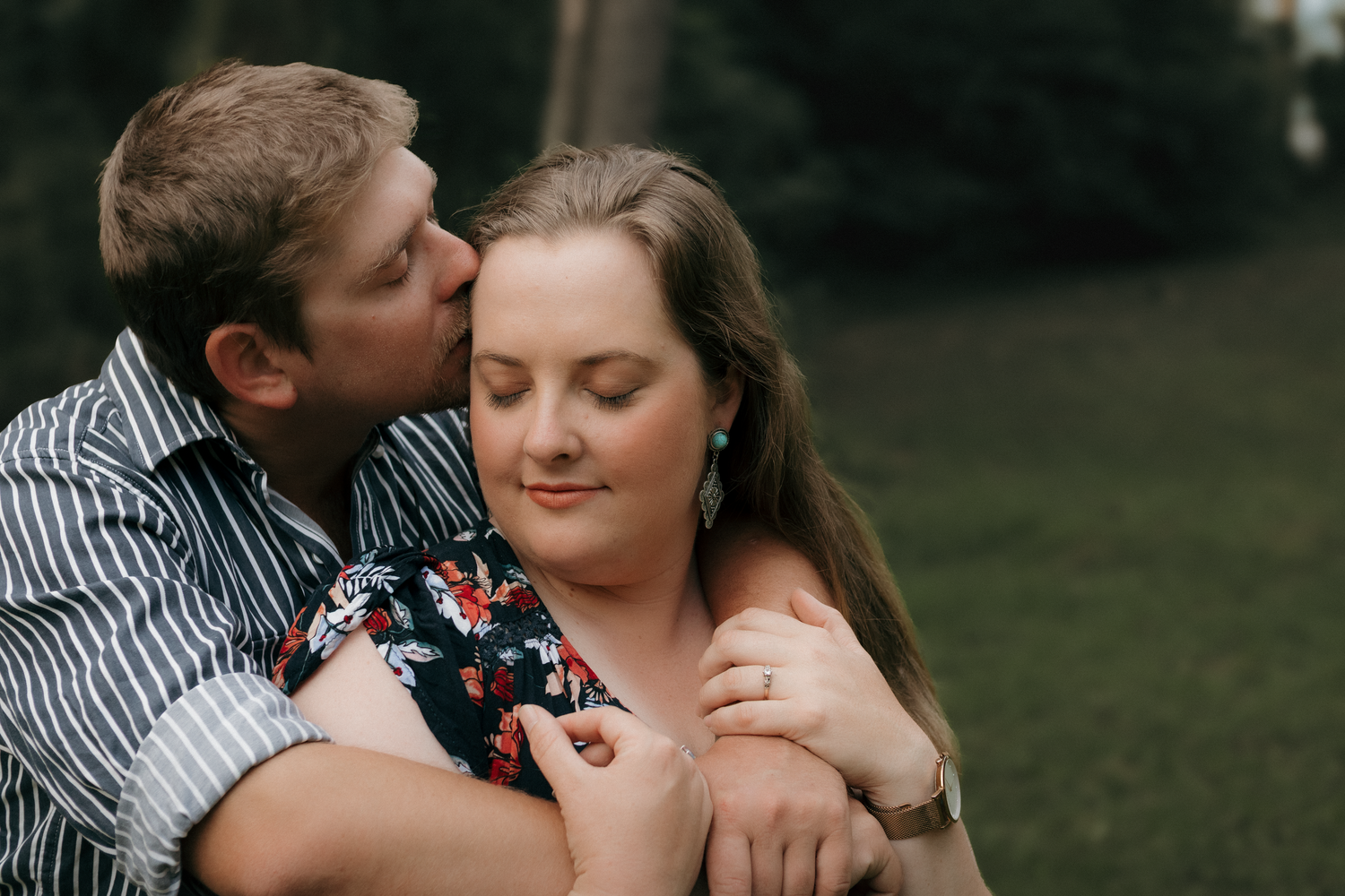 Couple embracing outdoors, man kissing woman on cheek while hugging her from behind.