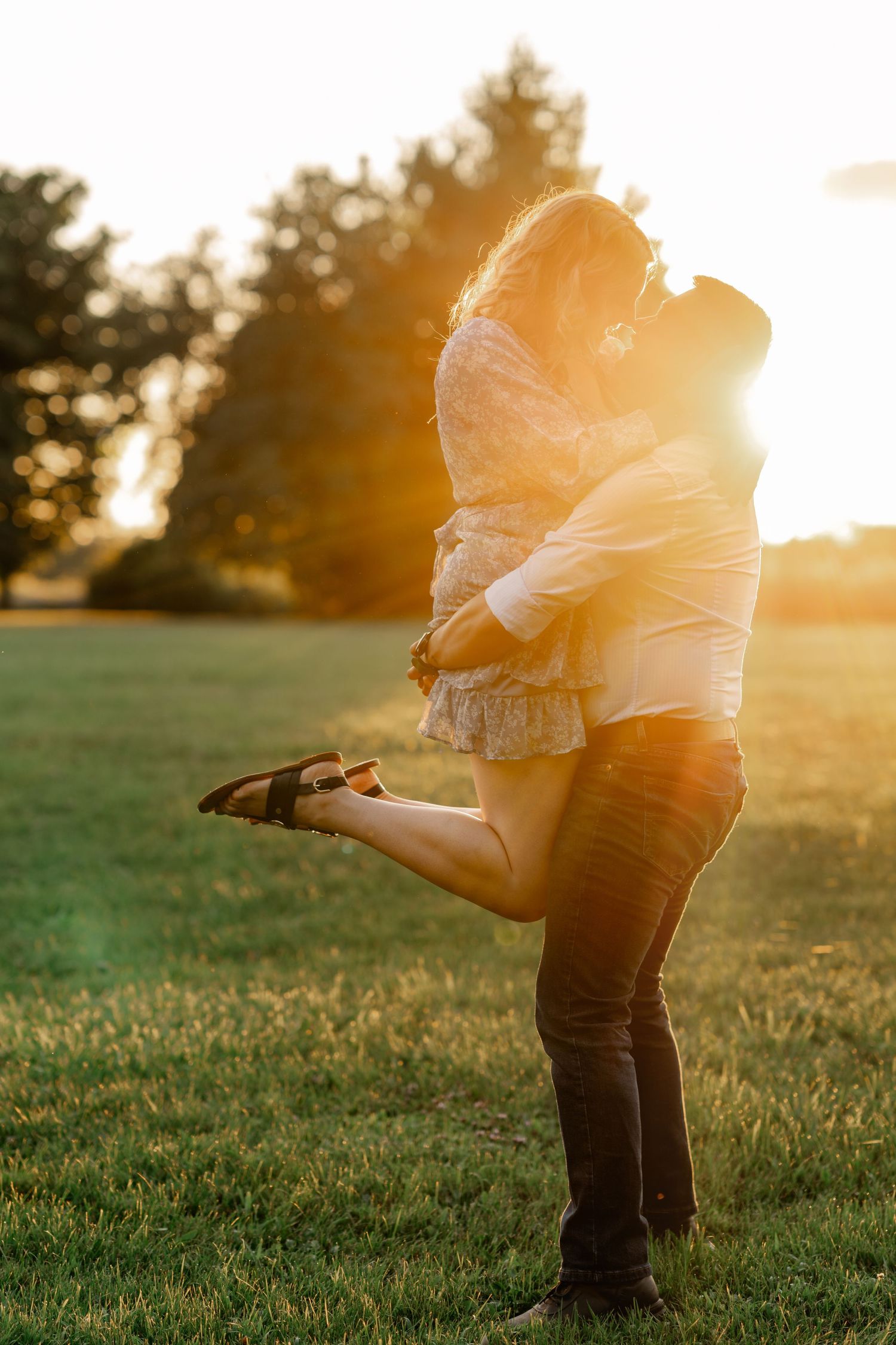 A couple embraces in a romantic sunset moment on a grassy hillside with golden sunlight streaming through their silhouettes.