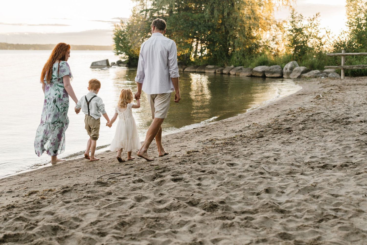 Family of four walking hand-in-hand along a sandy beach shore at sunset near calm water.