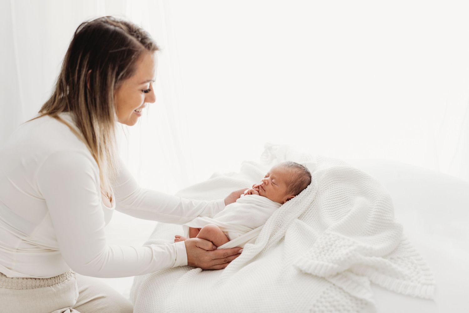 A person in white sits on a bed holding a newborn baby wrapped in soft white blankets.