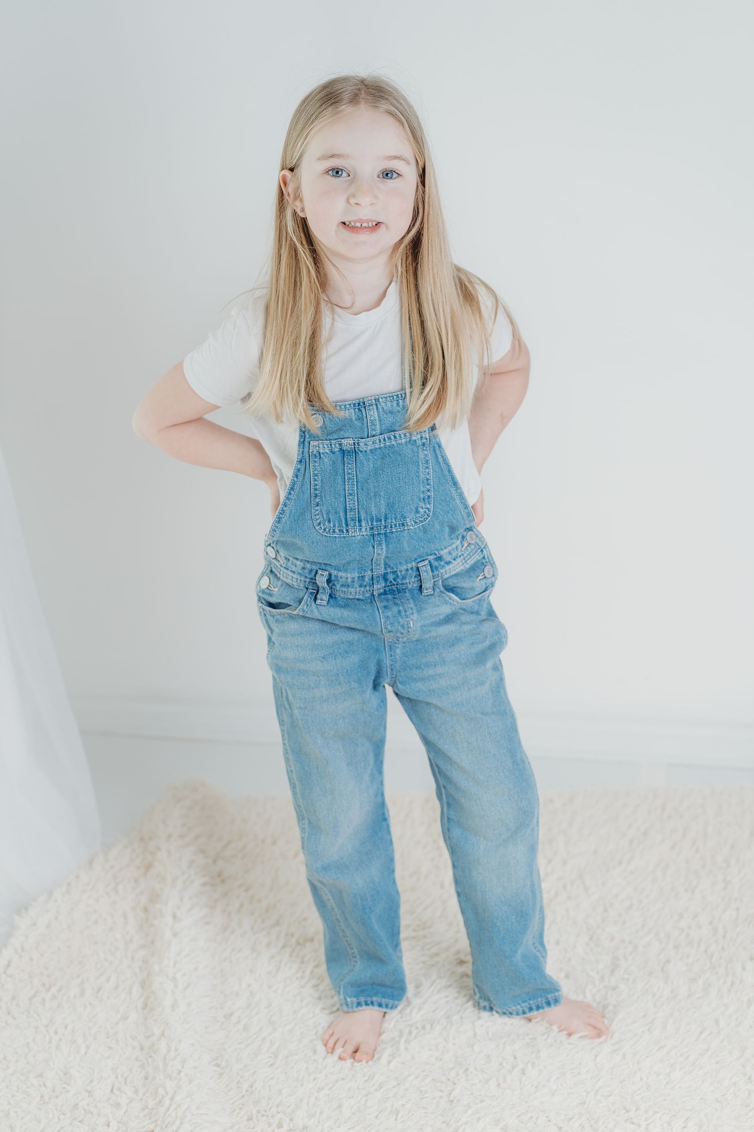 Three progressive photos show a toddler in denim overalls playing and posing on a white carpet.