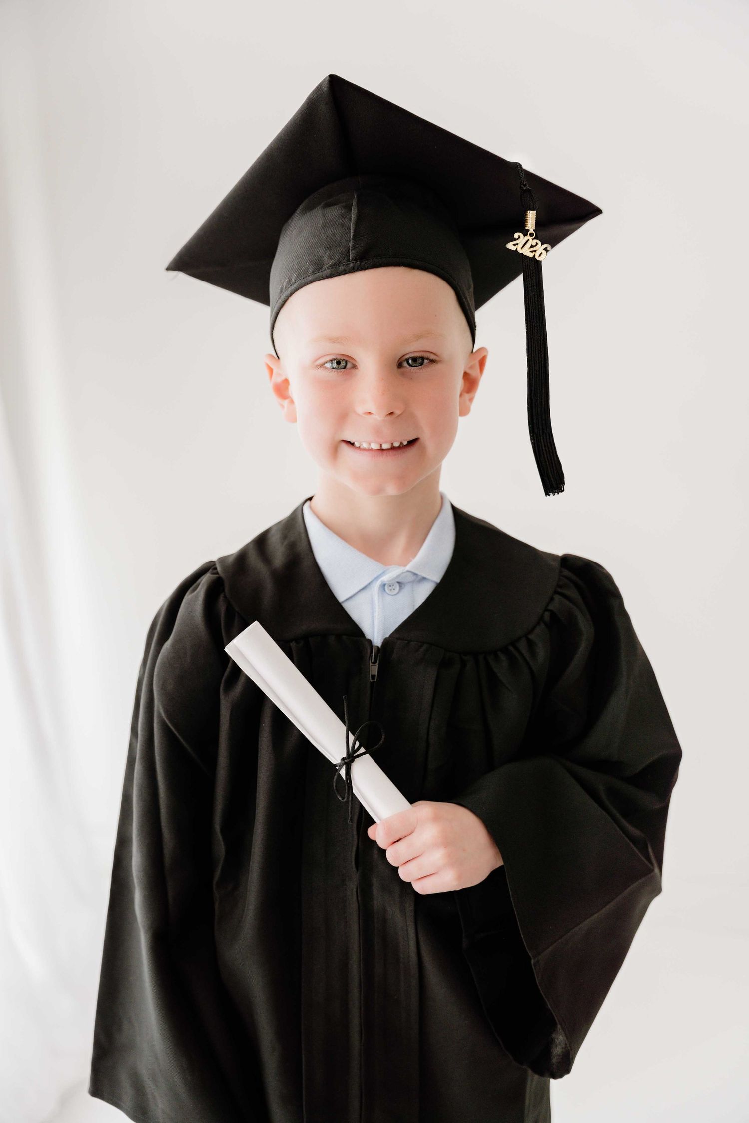 Young boy in black graduation cap and gown smiling while holding a diploma on white background.